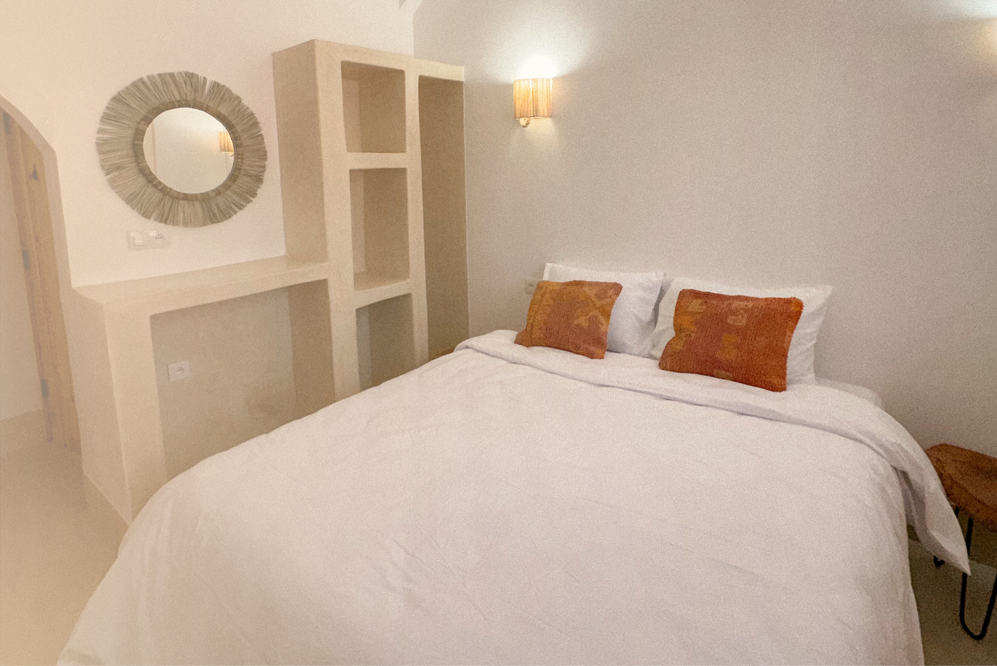 A neatly made bed with white sheets and two orange throw pillows in a minimalist hotel room. There is a round mirror and a small bookshelf on the wall, along with a wall-mounted sconce providing warm lighting.