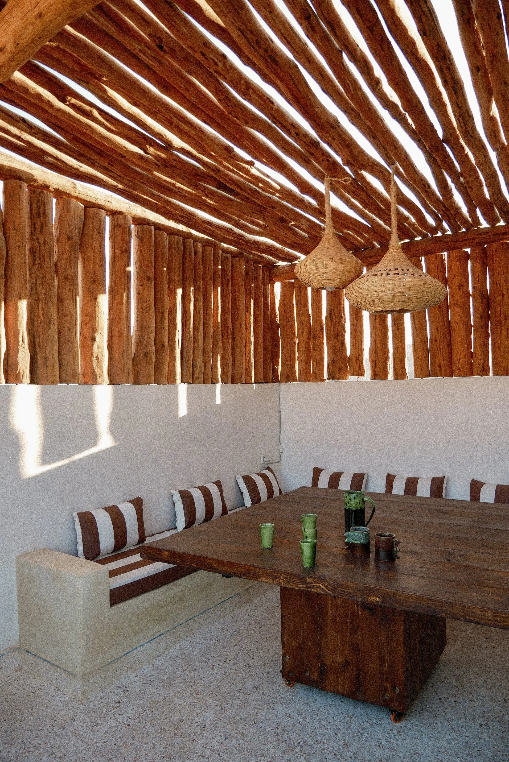 A rustic dining area with a wooden table, a built-in bench with striped cushions, and hanging woven pendant lights. The space features a wooden slat ceiling and a wall of vertical wooden slats allowing sunlight to filter through.