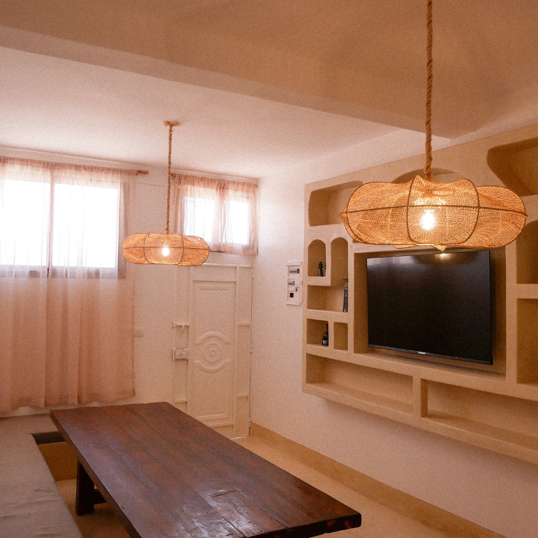 Living room with two hanging wicker pendant lights, a flat-screen TV on a built-in wall shelf, a wooden table, and light-colored curtains on windows.