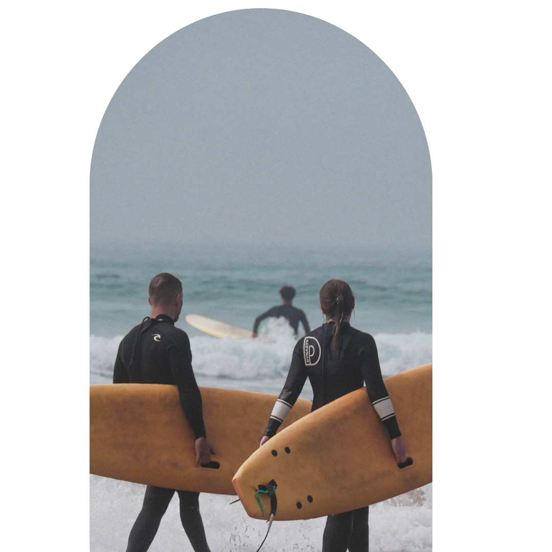Two surfers walking on the beach holding surfboards with their backs to the camera, heading towards the ocean with another surfer in the water.