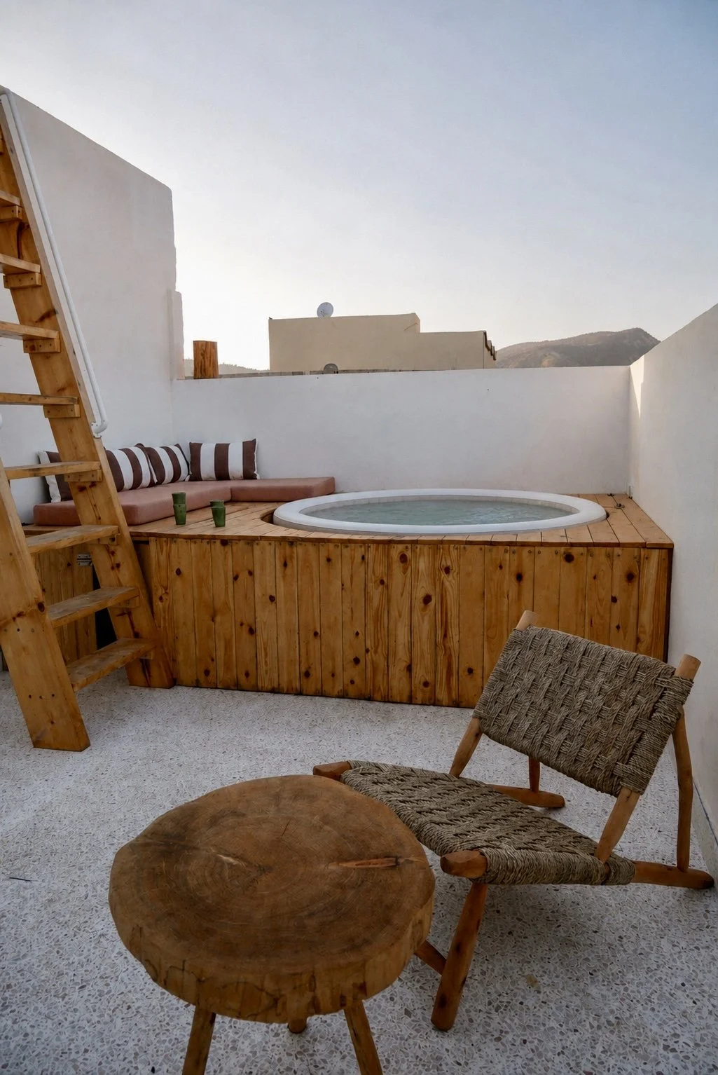 Rooftop terrace with hot tub, wooden seating area with striped cushions, woven chair, and a small round wooden table, against white walls and a clear sky.