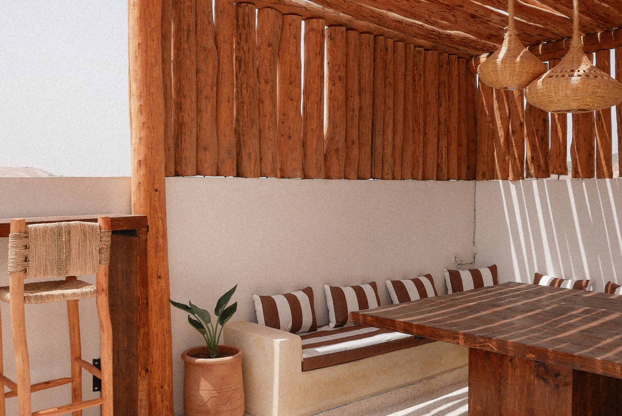 Rustic indoor space with wooden furniture, hanging wicker lamps, a striped cushioned bench, a tall plant in a terracotta pot, and a wooden chair, all illuminated by sunlight with shadows on the wall and floor.