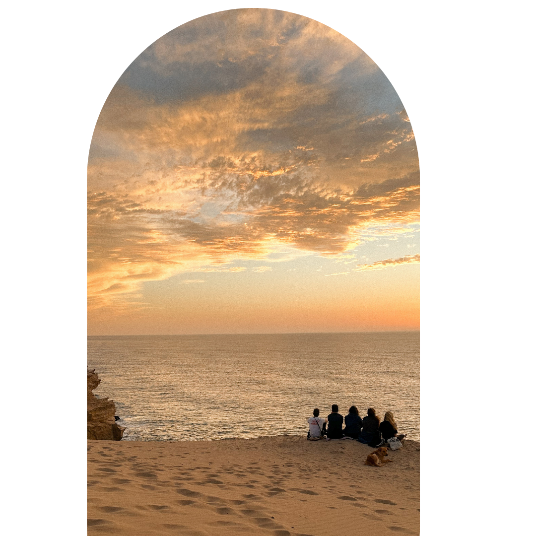 People sitting on a sandy beach watching a sunset over the ocean, with clouds in the sky.