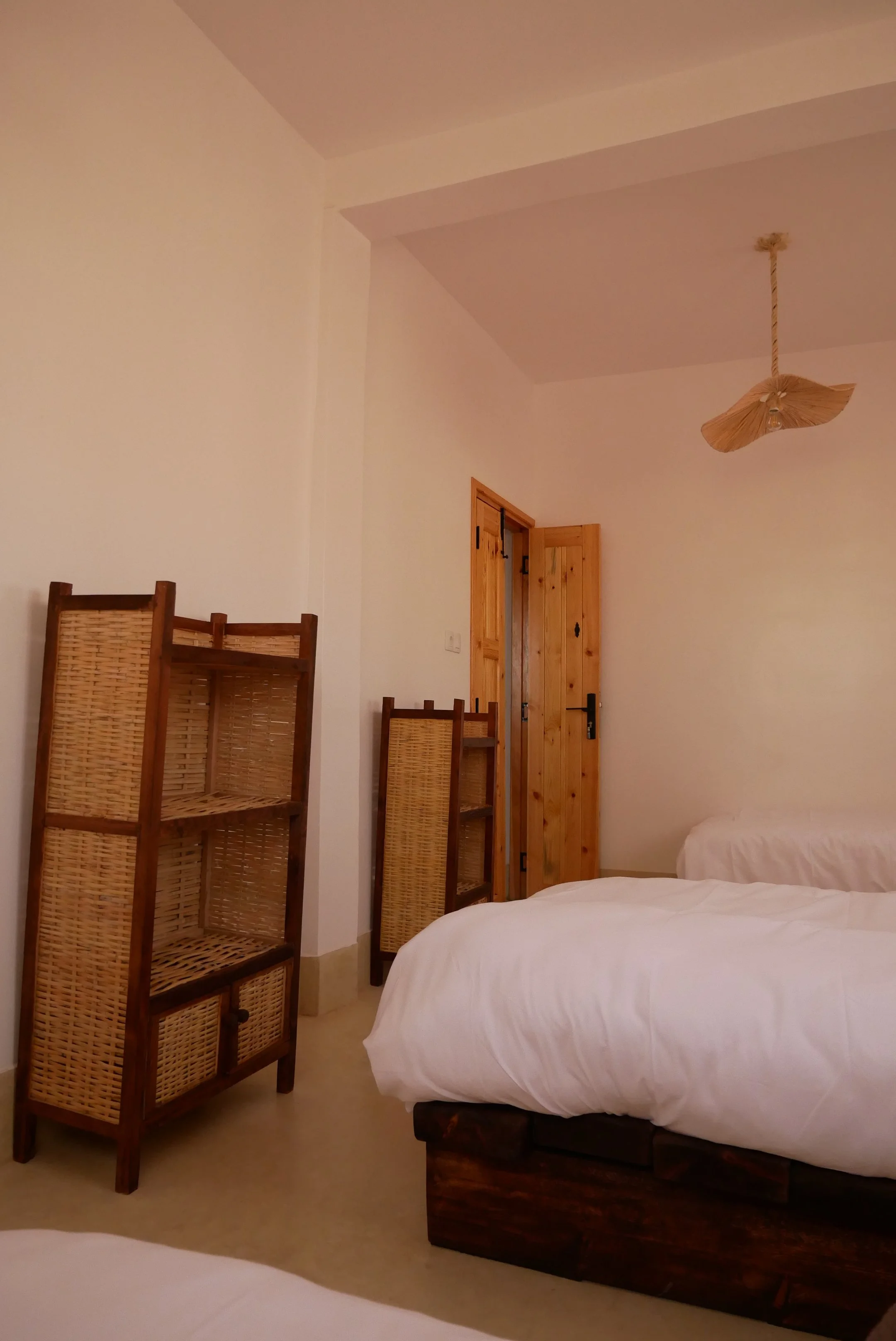A bedroom with two beds, a wooden door, a woven wood lamp hanging from the ceiling, and four wooden chairs stacked against the wall.