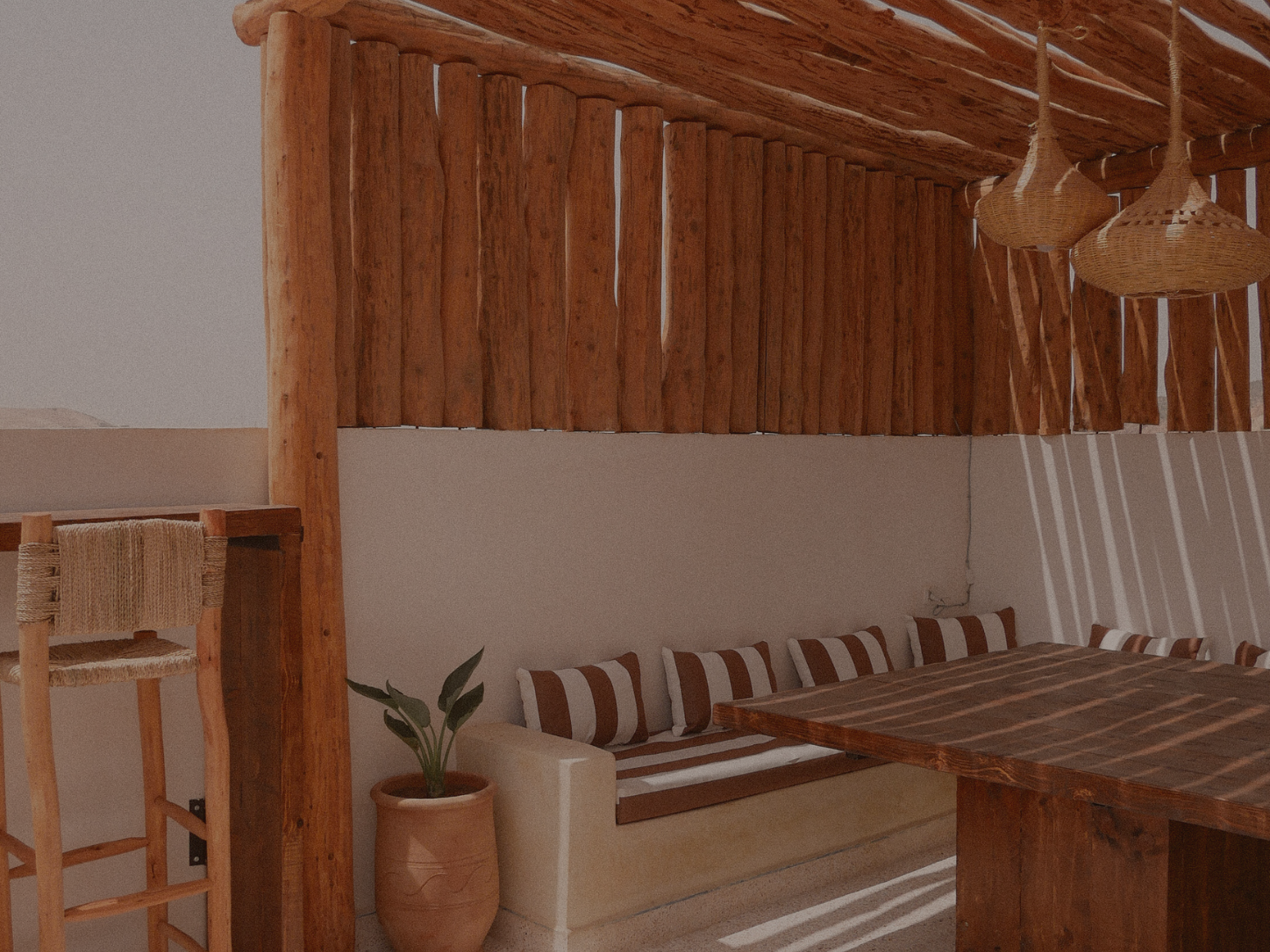 Interior view of a room with wooden and white decor, a built-in bench with striped cushions, a wooden table, hanging woven pendant lights, and a potted plant