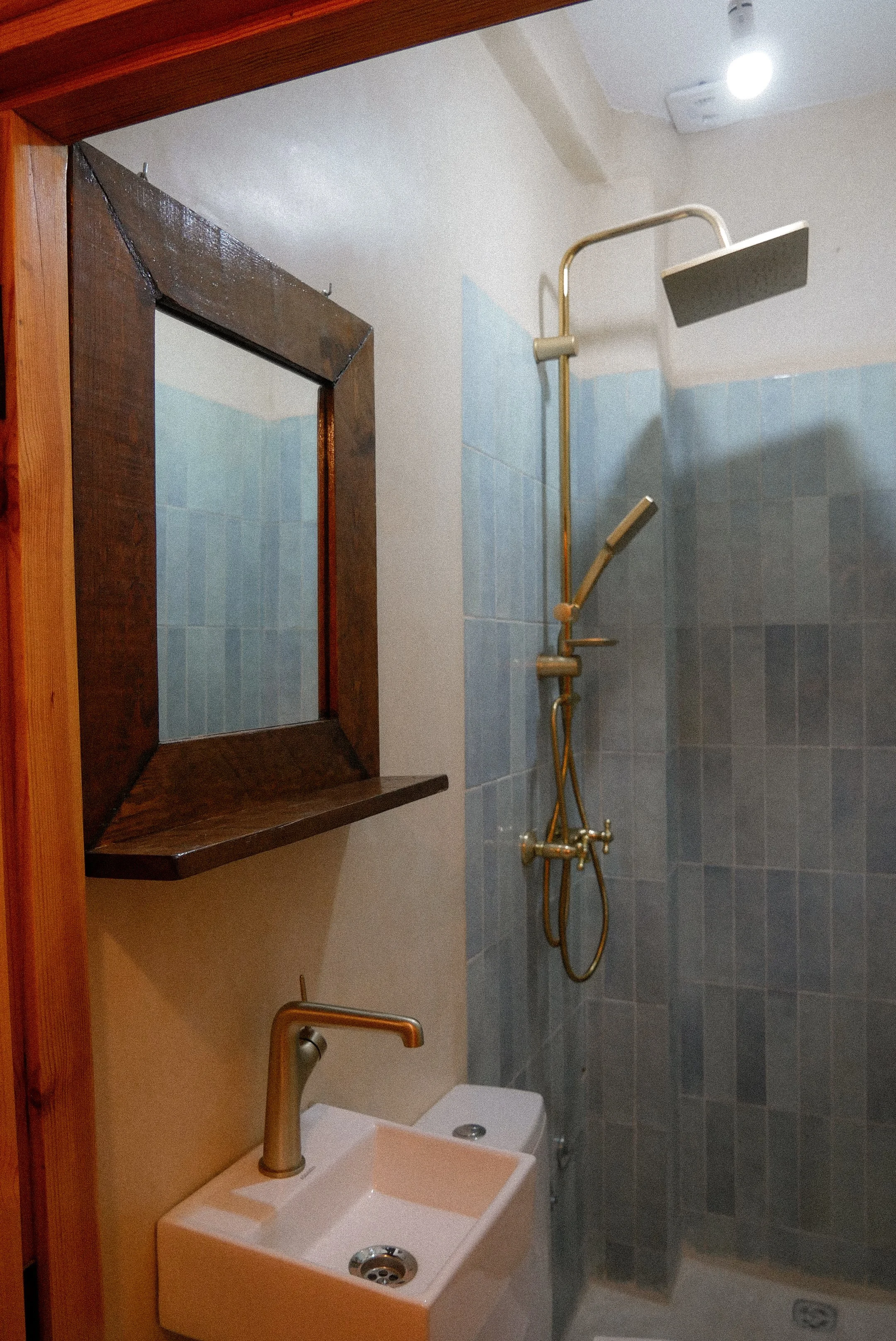 A small bathroom with a blue tiled shower area, a wall-mounted shower head, a white sink with a gold faucet, a wooden-framed mirror, and a small white toilet. The bathroom appears clean and organized.