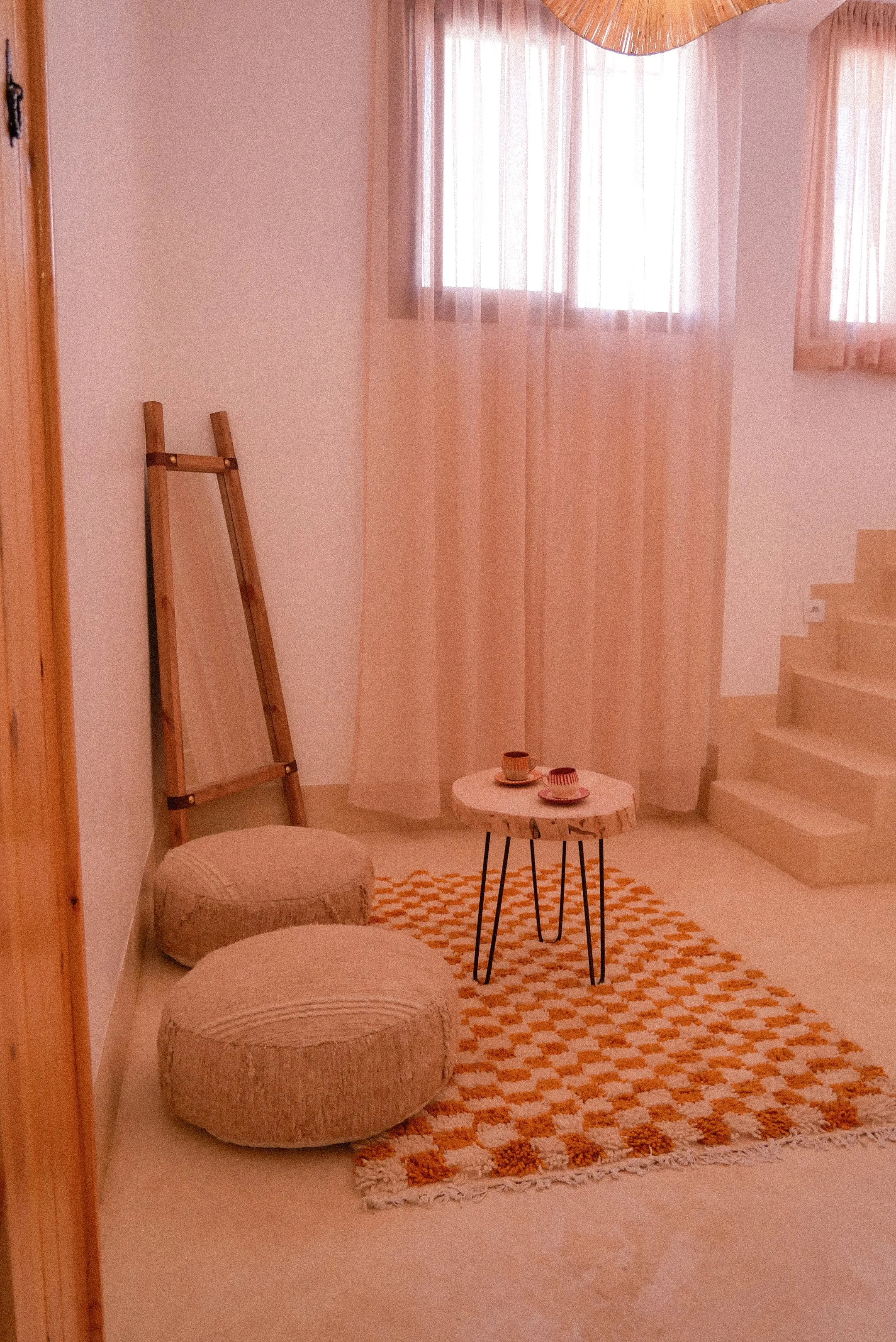a cozy living room corner with two round beige poufs, a small white coffee table with two cups, a patterned rug, sheer curtains, a wooden ladder leaning against the wall, and a staircase with white steps to the right