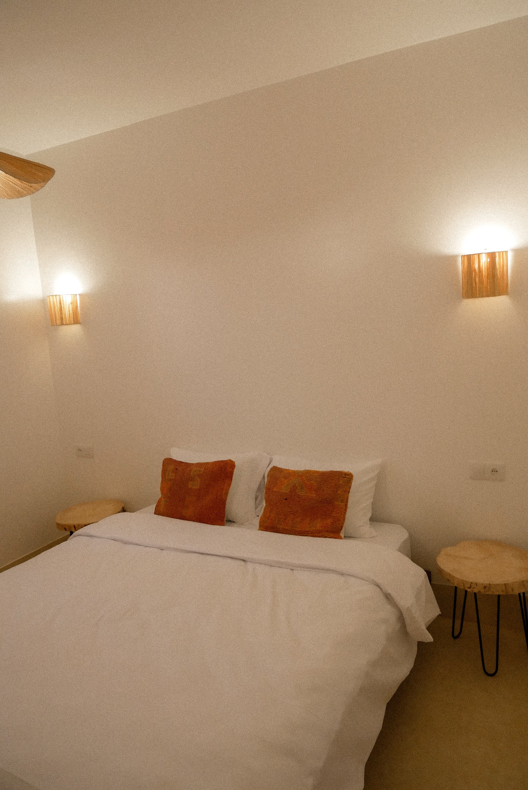 A neatly made bed with white linens, decorated with three orange and red pillows, flanked by two round wooden side tables with black legs, and illuminated wall-mounted lights above.