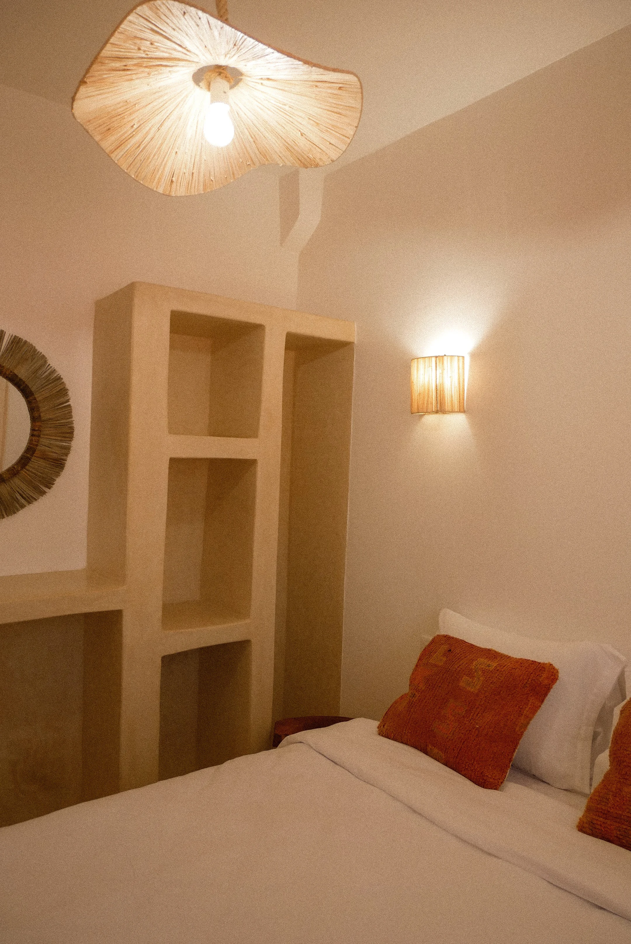 A bedroom with a white bed, two orange pillows, a wall-mounted light, a ceiling light with a beige lampshade, and an empty beige bookshelf.