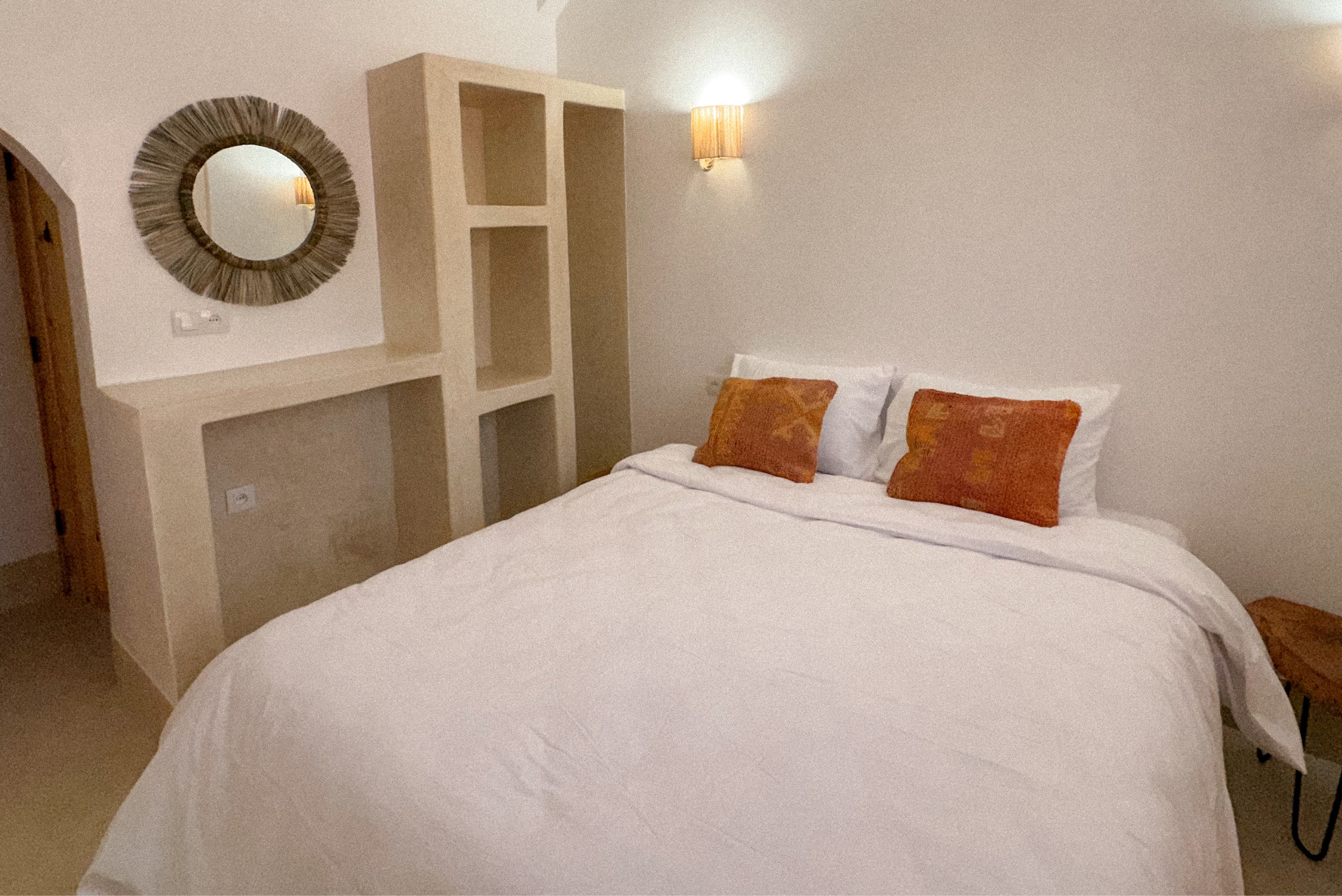 A neatly made bed with white sheets and two orange throw pillows in a cozy bedroom with beige walls, a wall-mounted lamp, a mirror with a textured frame, and built-in shelving.