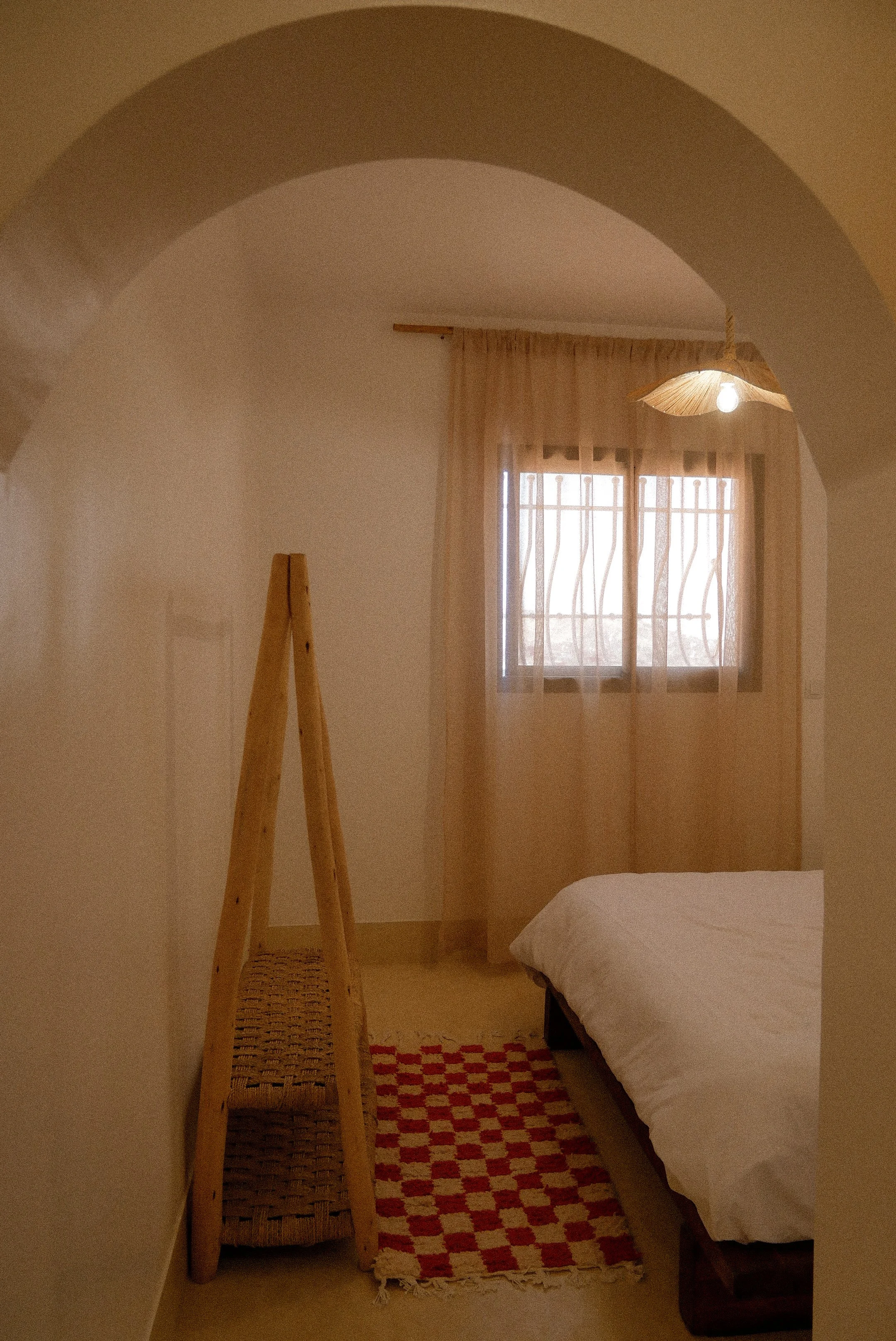 A small, cozy bedroom with a bed covered in white sheets, a window with light curtains, a wooden ladder, and a patterned rug on the floor. There is a woven ceiling lampshade and natural light coming through the window.