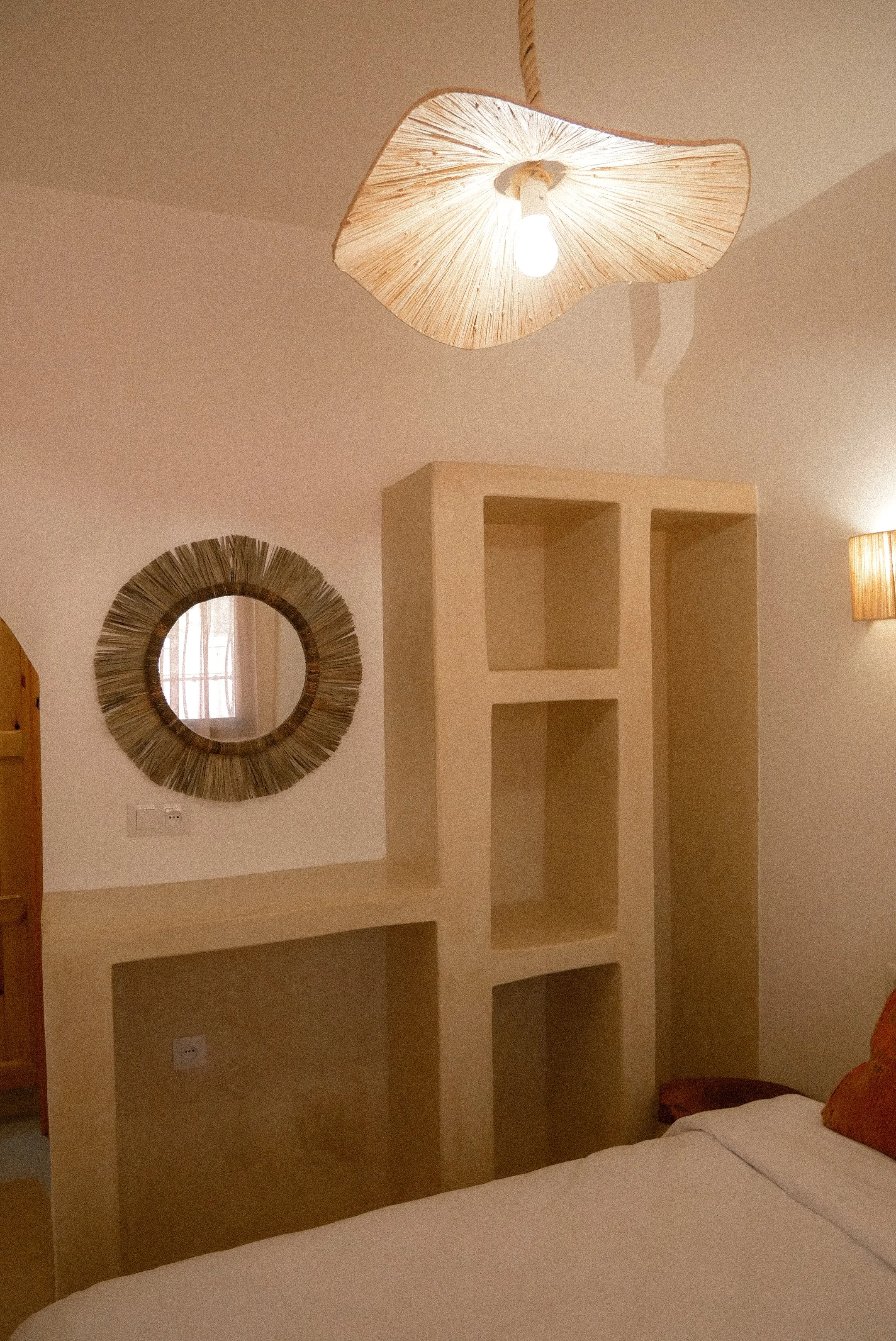 Interior of a bedroom featuring a bed with white bedding in the foreground, a built-in beige bookshelf unit along the wall, a round mirror with a wooden frame on the wall, a window with curtains reflected in the mirror, and a hanging wooden lampshade with a light bulb.