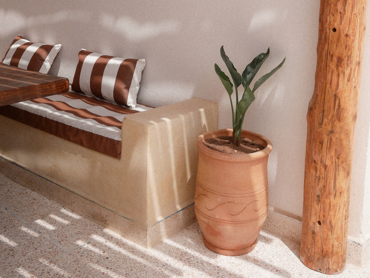 A beige stone bench with two brown and white striped pillows and a brown and white striped runner, next to a tall terracotta plant pot with a green plant, and a large wooden log leaning against the wall. Sunlight creates shadows on the floor.