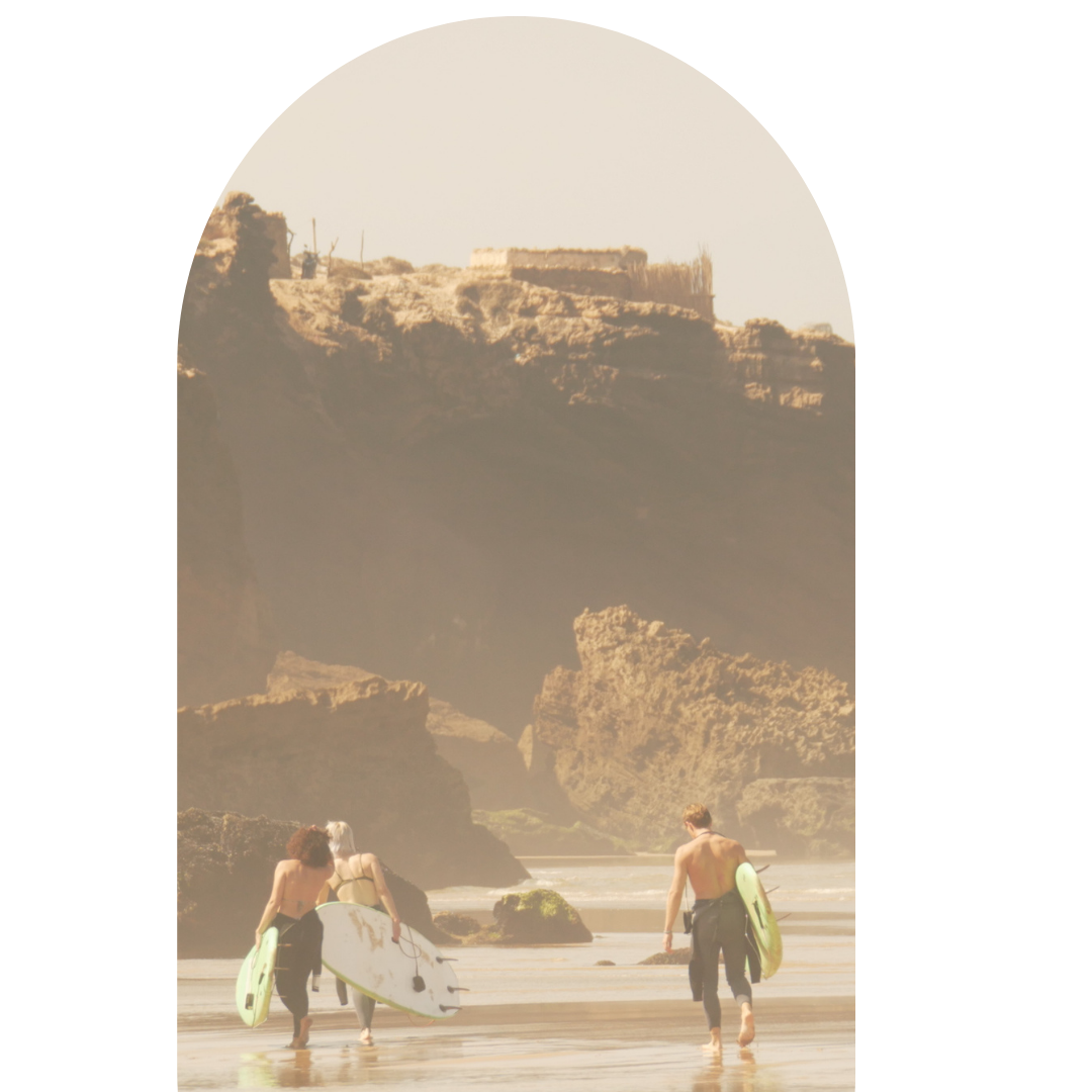 Three surfers walking on the beach carrying surfboards, with rocky cliffs and a large arch in the background.