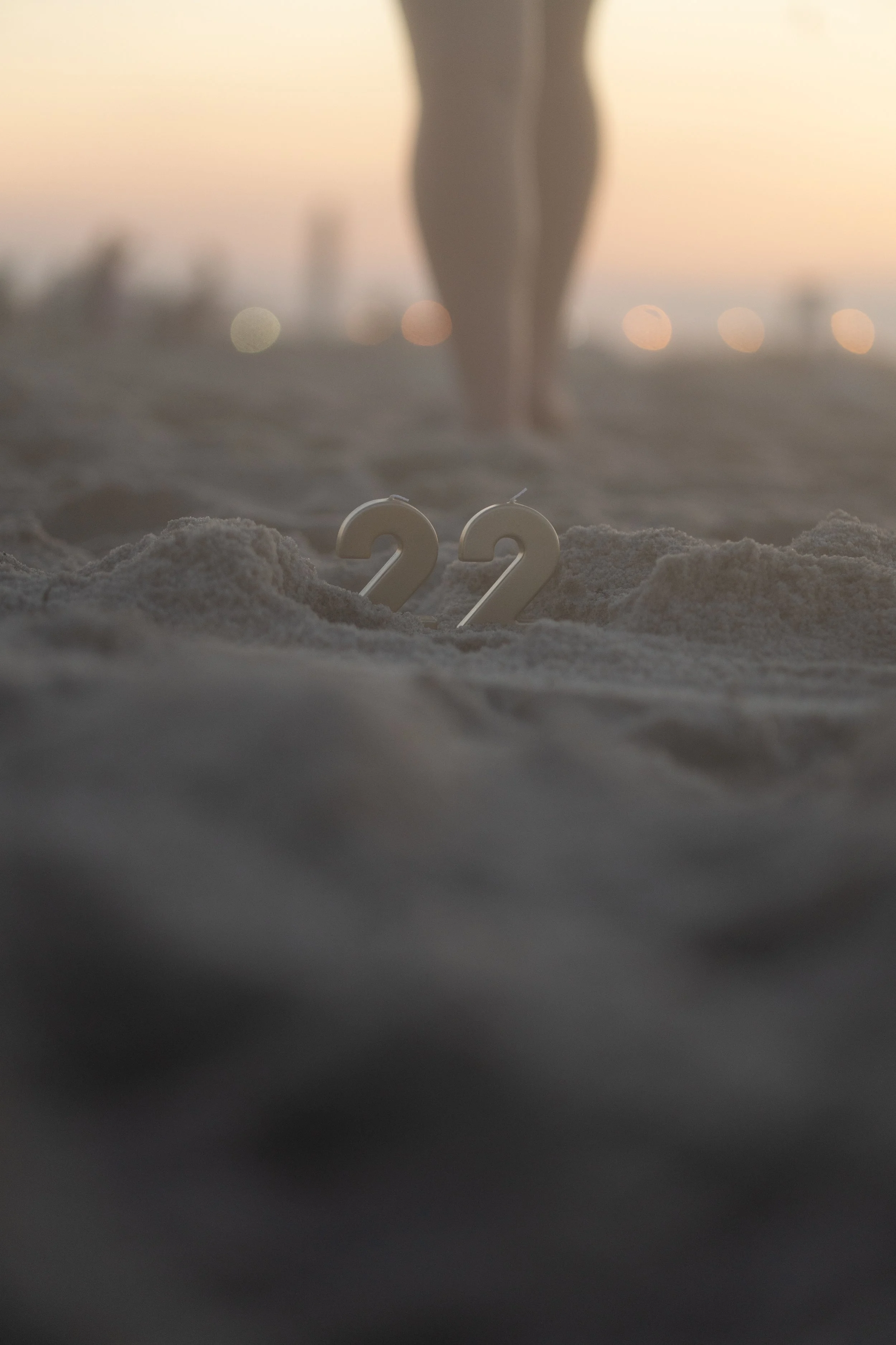Two white candles shaped as the numbers 2 and 2 are placed in the sand on a beach during sunset, with a blurred human figure standing in the background.