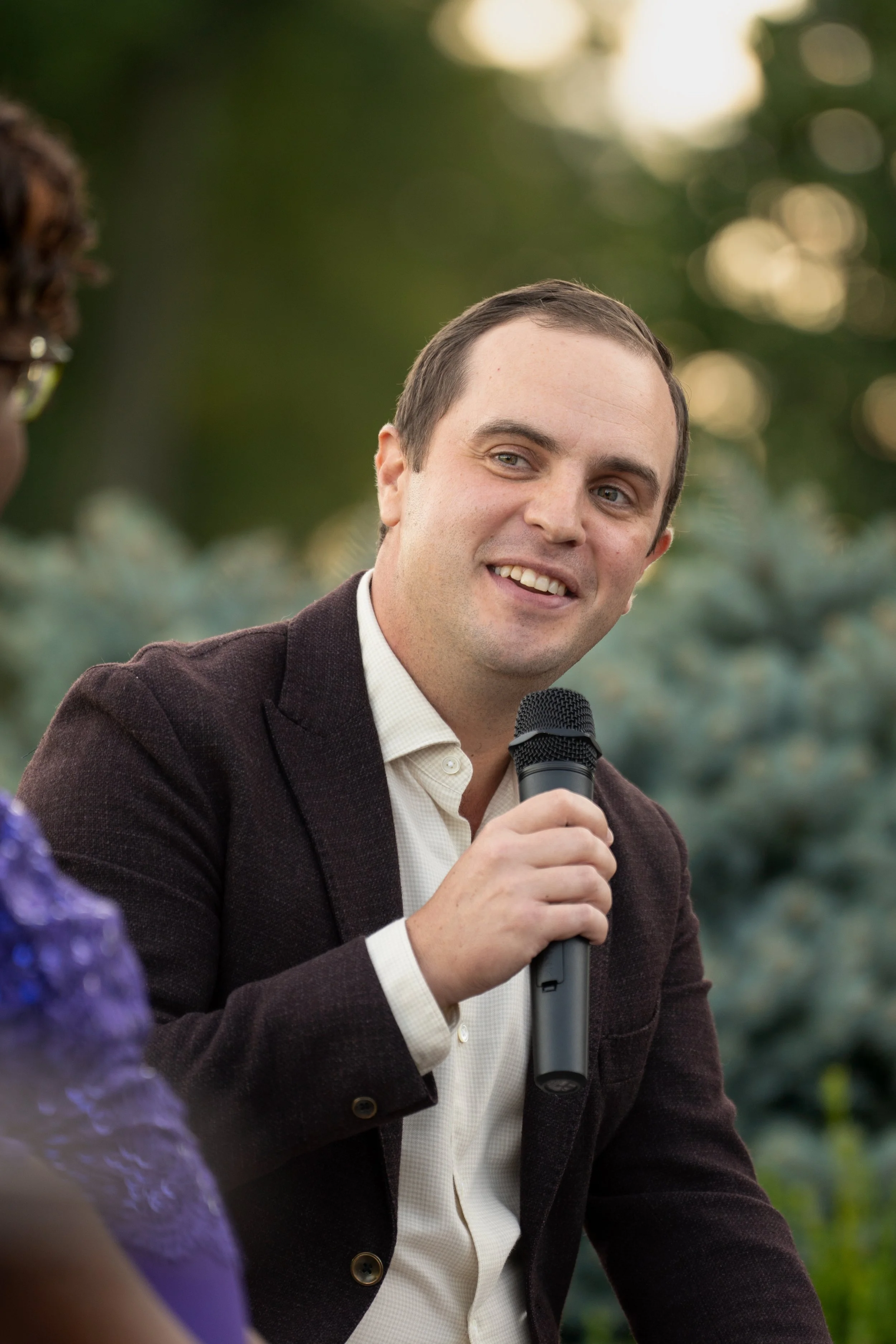 A man with short brown hair, wearing a dark blazer and white shirt, holding a microphone and smiling during an outdoor event with blurred green trees in the background.