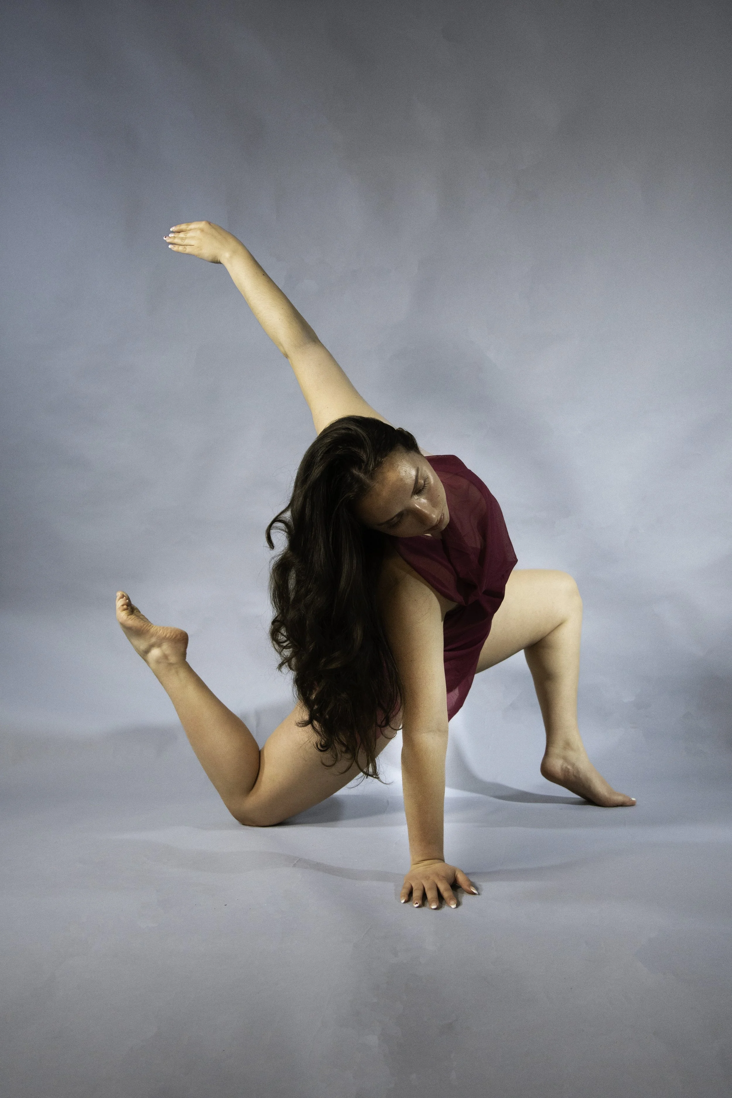 Two women practicing yoga together, one woman supporting the other in a balancing pose, on a plain gray backdrop.