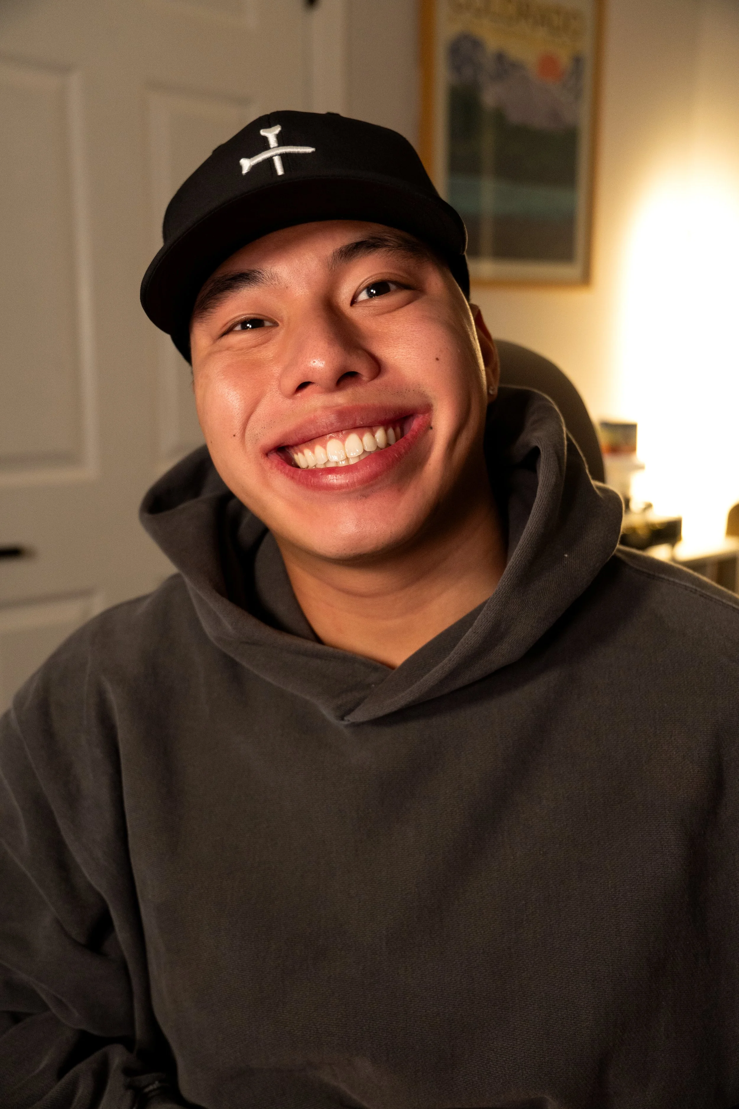 A smiling young man wearing a black baseball cap with a white logo and a dark gray hoodie, sitting in a warmly lit room with a framed picture on the wall behind him.