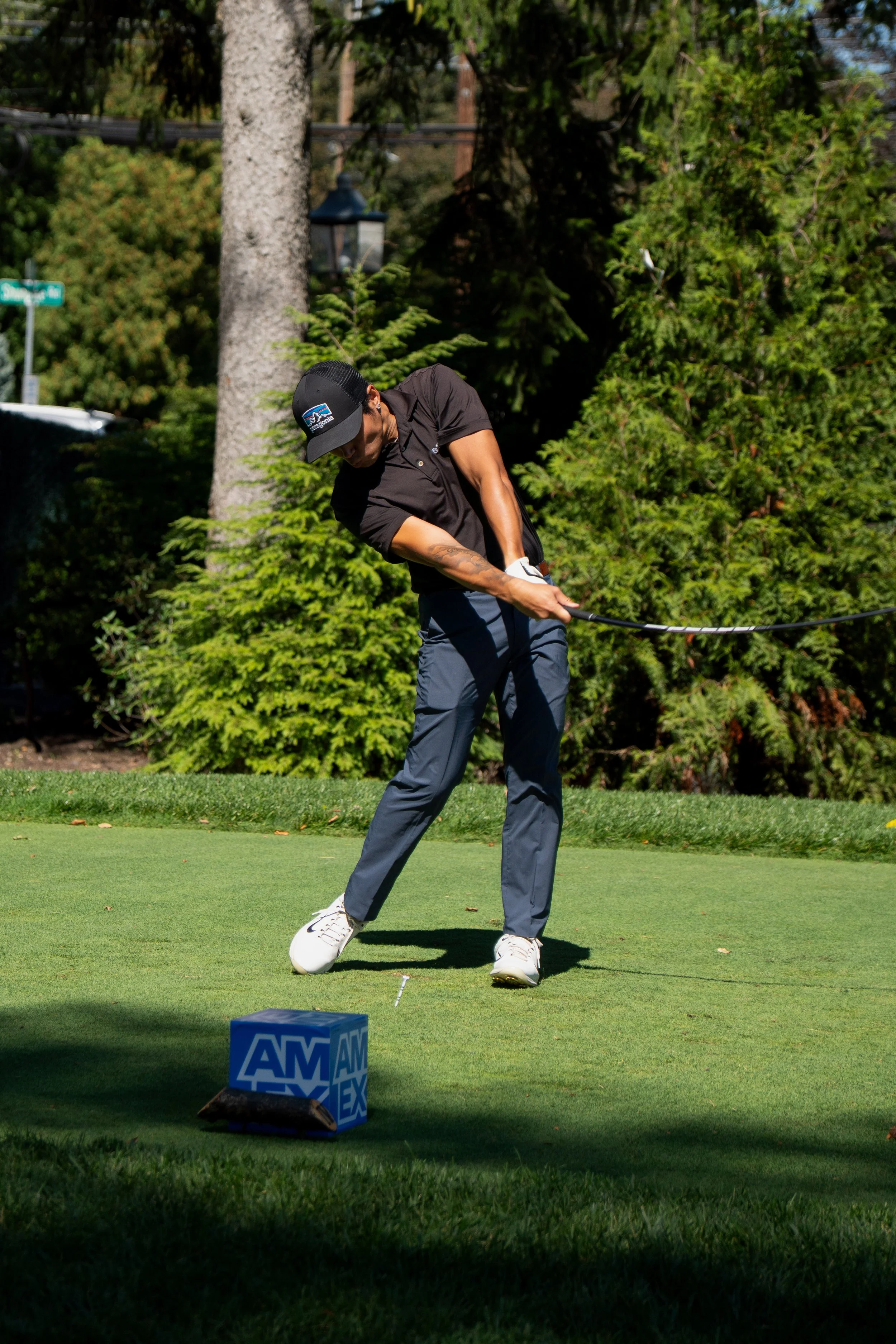 A man playing golf, preparing to hit the ball on the green, with focus on his stance and swing. He is wearing a black cap, black shirt, gray pants, and white golf shoes. There are trees and a street sign in the background, and a blue box with 'AM' partially visible in the foreground.