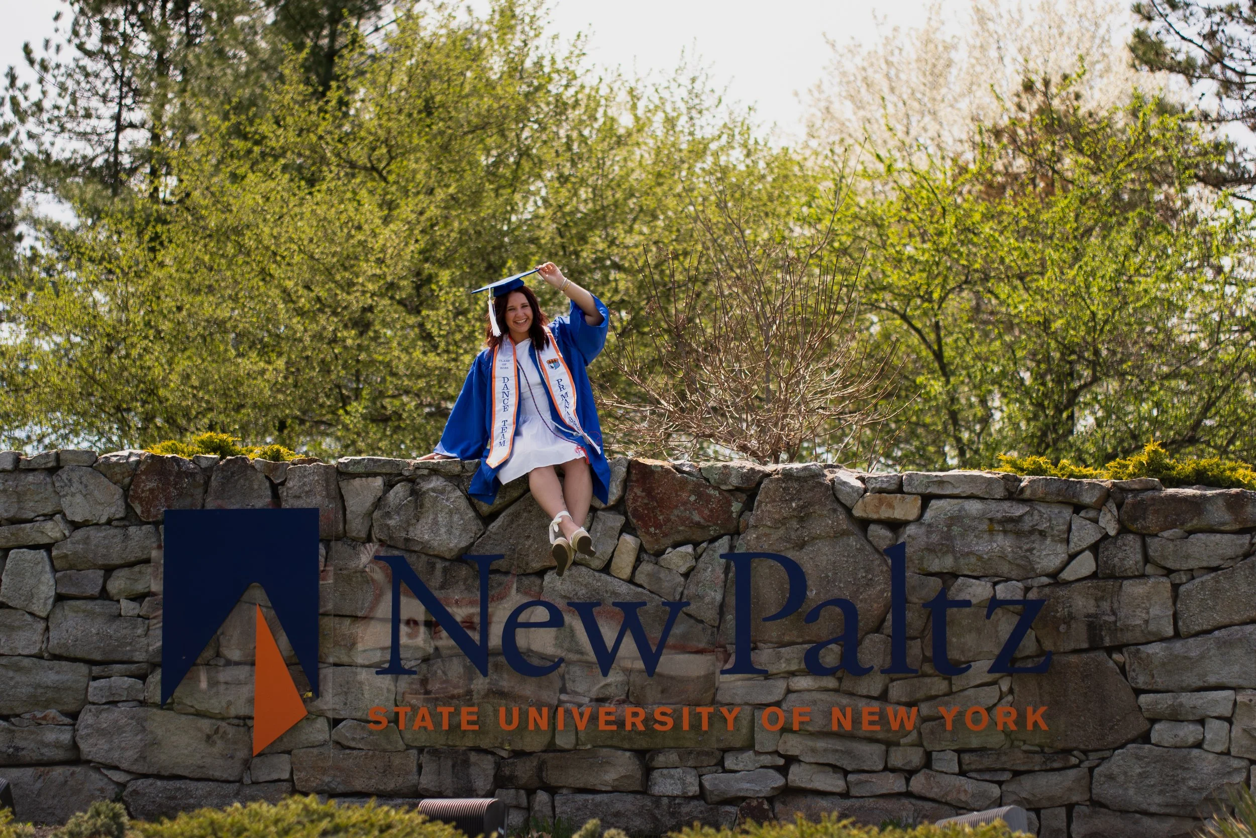 Smiling young woman in a blue graduation gown and cap sitting on a stone wall in front of a sign that reads "State University of New York at New Paltz" with trees and greenery in the background.