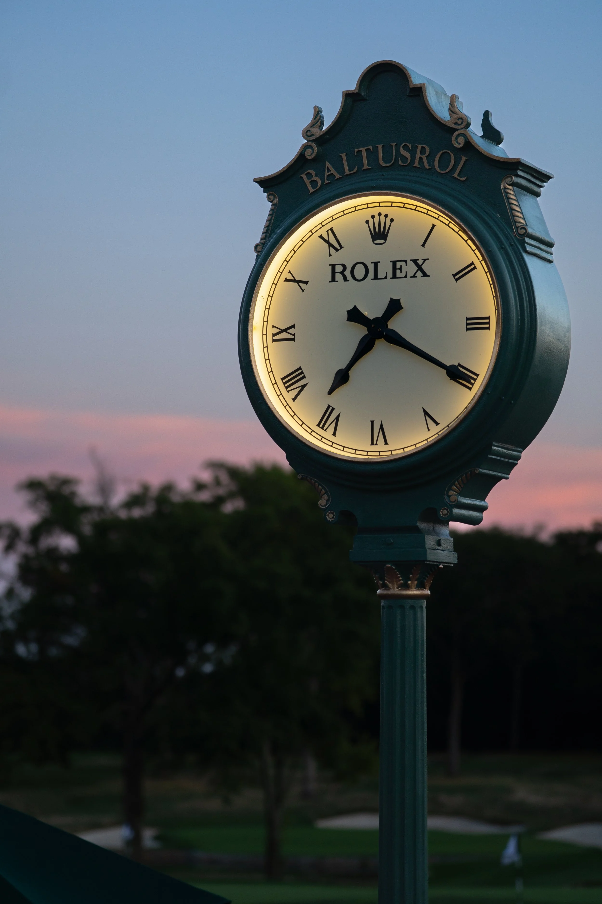 A vintage-style clock on a pole displaying the time as 2:19, illuminated against a dusk sky, with trees and a park visible in the background.