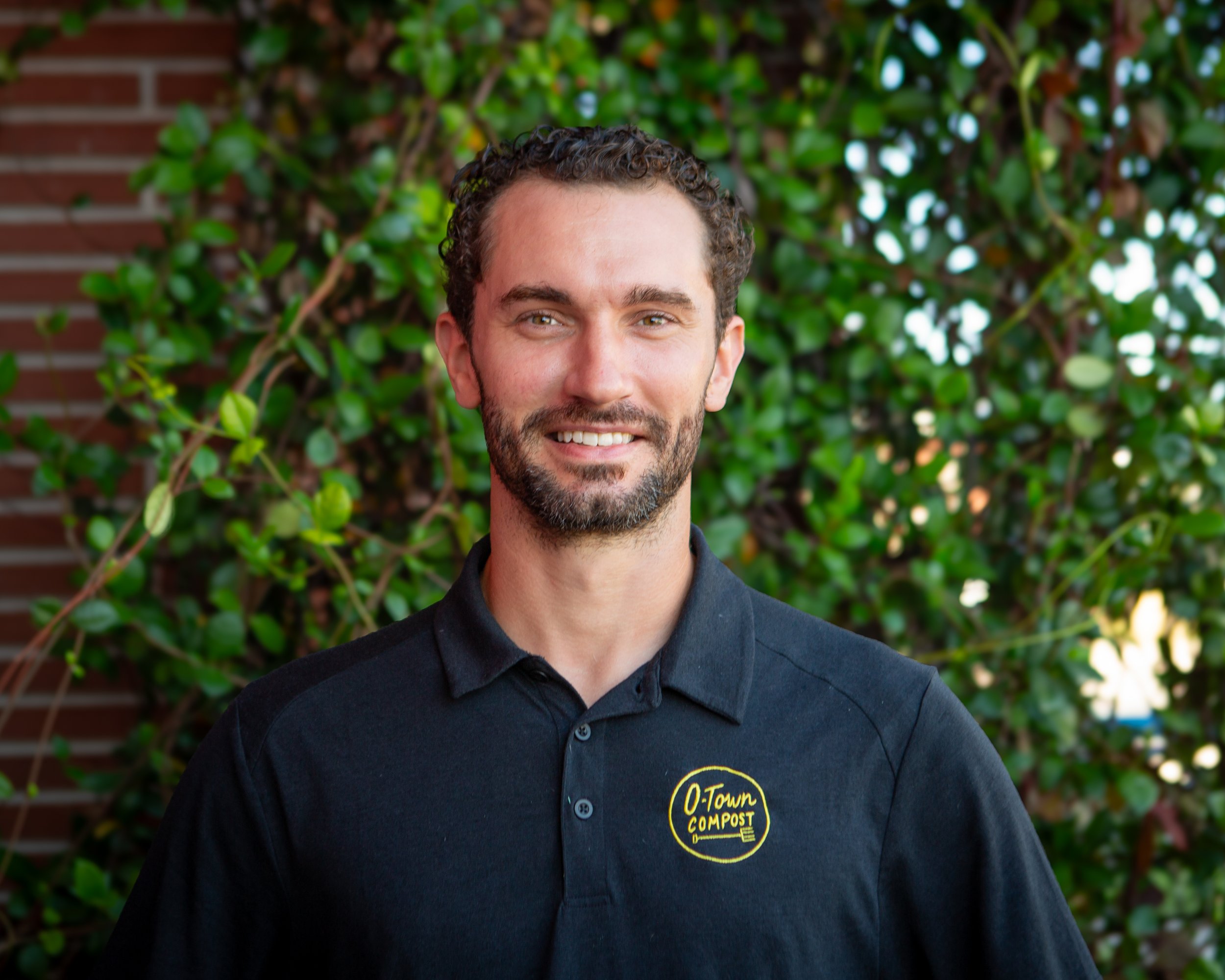 Close-up of a smiling man wearing a black O-Town Post Compost shirt, standing outdoors against a background of green leafy bushes and a brick wall.