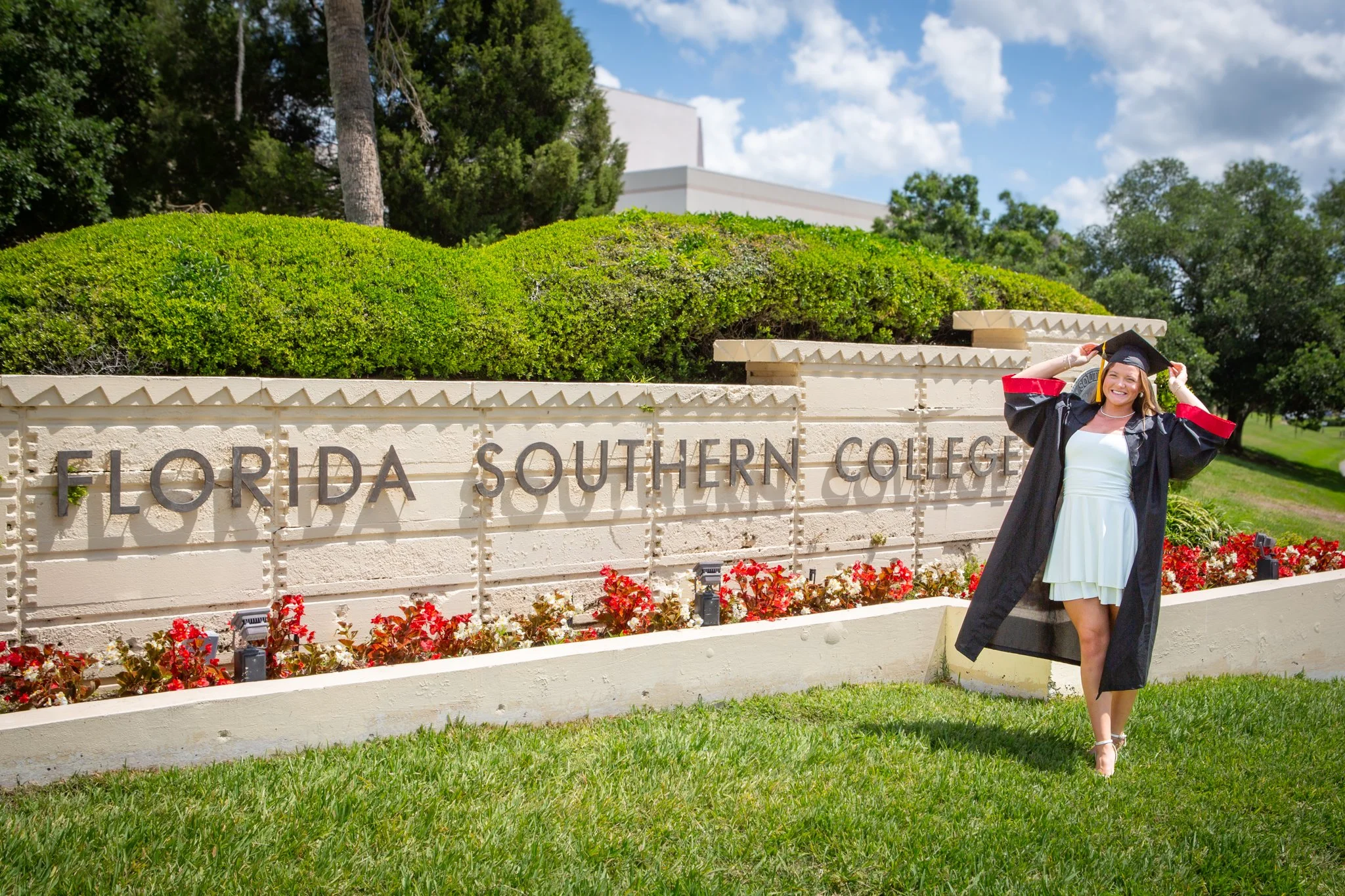 A woman in a graduation gown and cap standing outdoors in front of a cream-colored sign that reads 'FLORIDA SOUTHERN COLLEGE.' She is smiling and adjusting her cap with both hands. The background features green trees and a clear blue sky with some cl