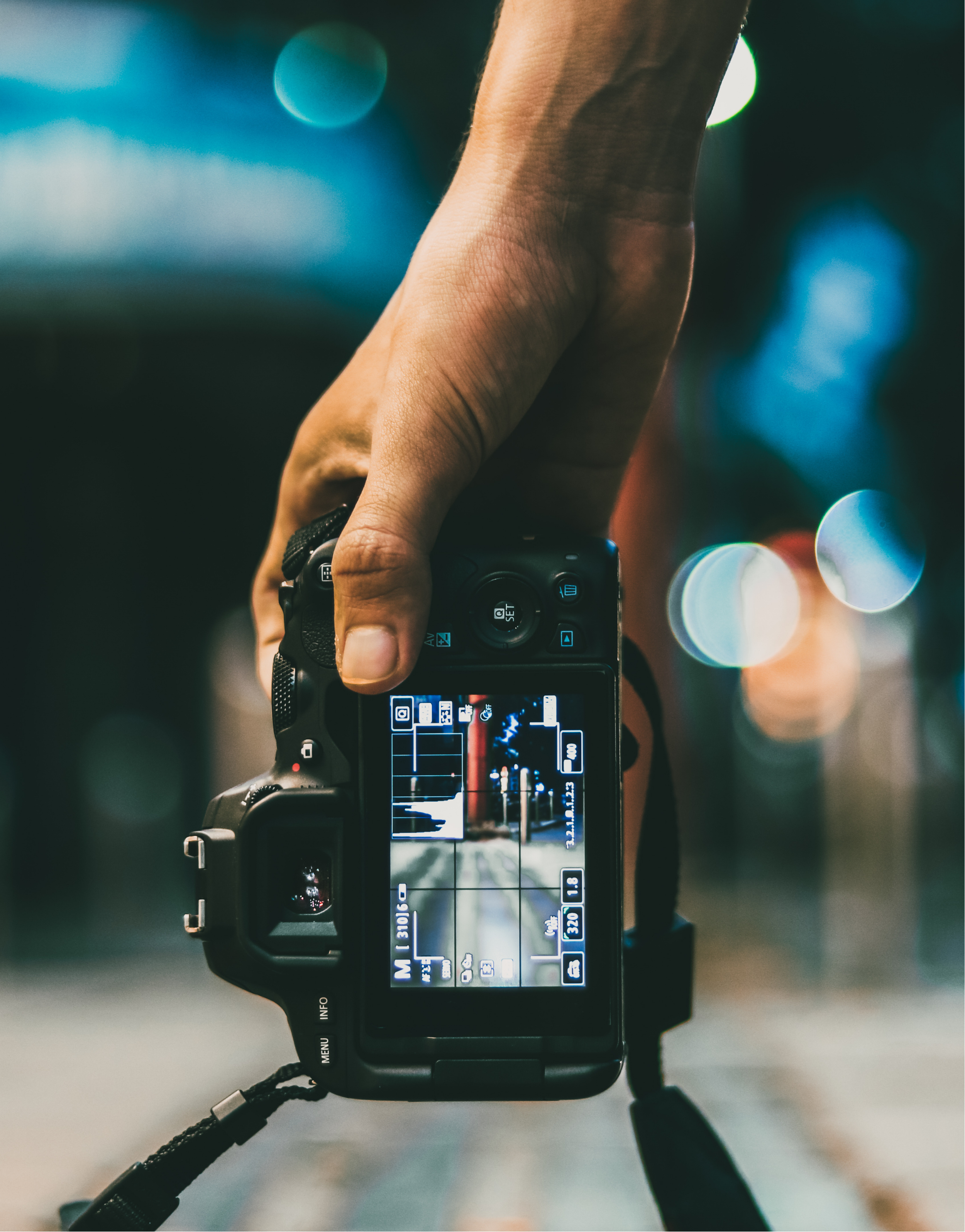 A digital camera with a wrist strap viewed from the back, displaying various settings on its screen, with a blurred background of city lights.