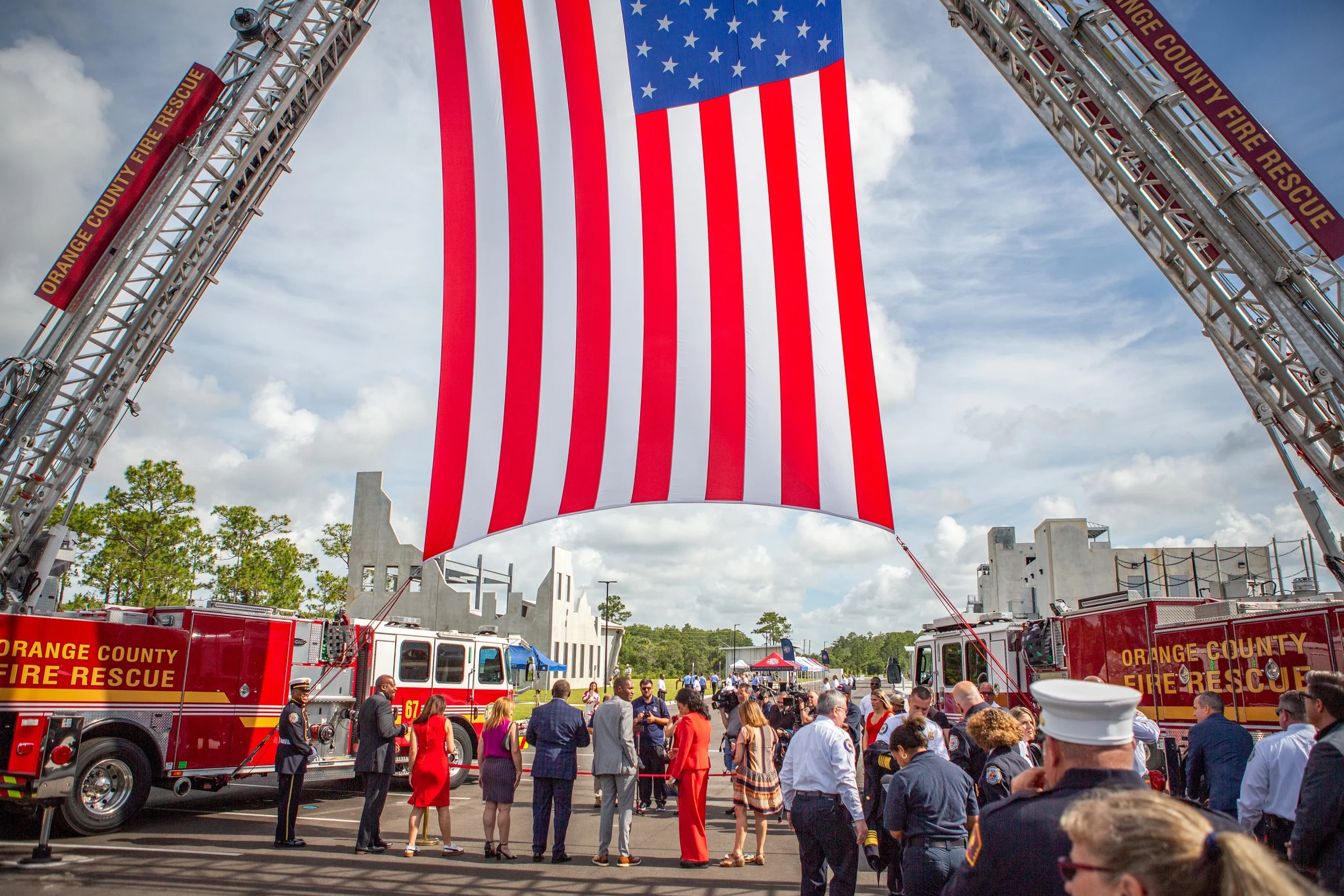 A large American flag hanging from a fire truck fire rescue ladder at an outdoor event with many people, including fire fighters, police officers, and civilians, gathered below.