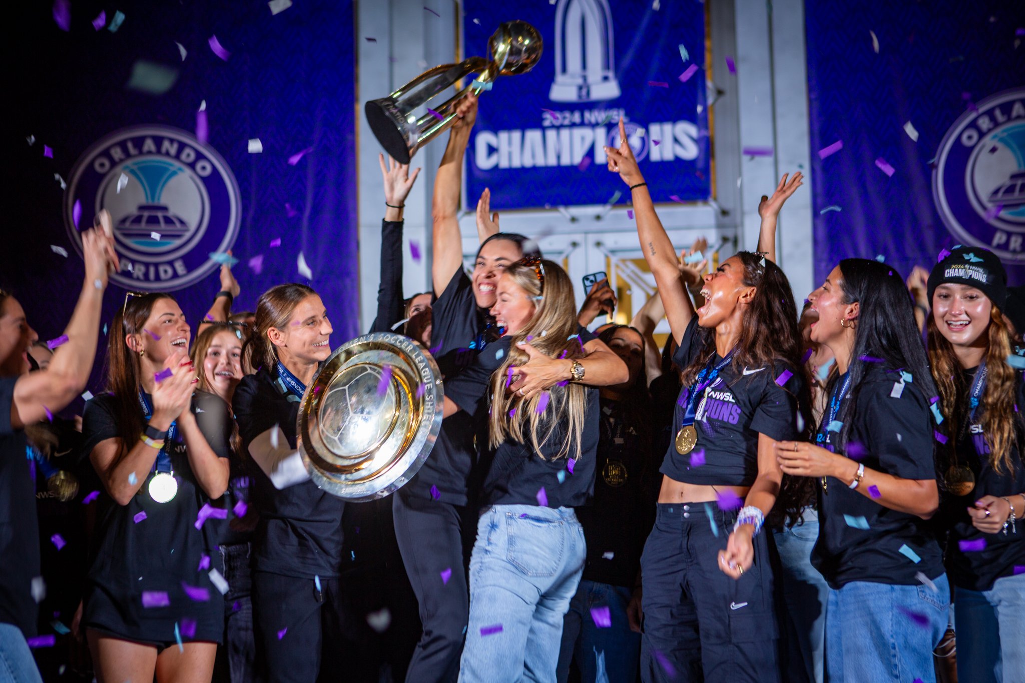 A group of women celebrating with confetti, holding a trophy, and wearing medals, at a championship event with a blue banner reading '2024 NWSL Champions' in the background.