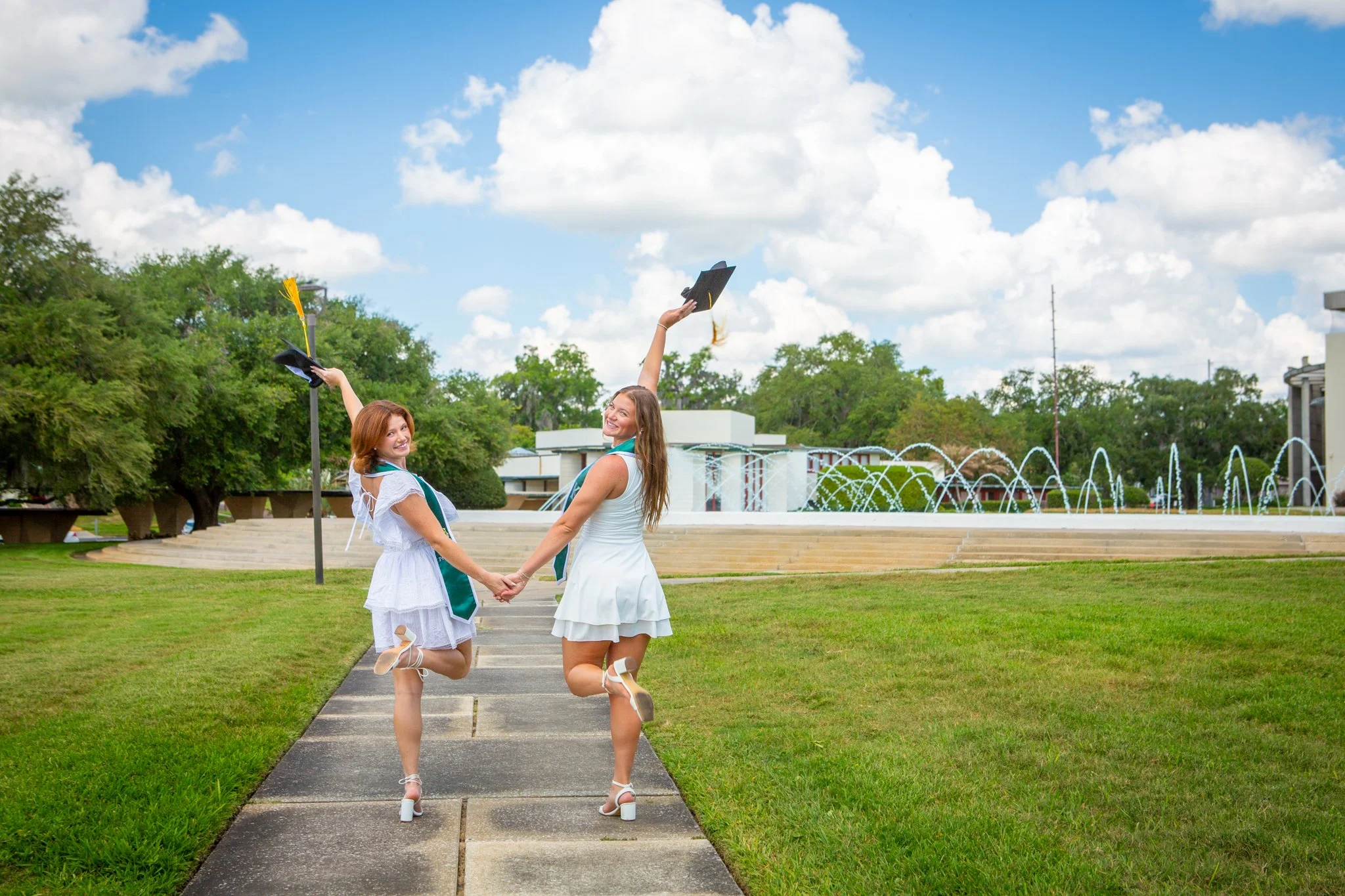 Two women in white dresses and high heels are holding hands and jumping on a paved walkway outdoors. They are wearing graduation caps with tassels. The background features a fountain, green trees, a blue sky with white clouds, and a building with a r