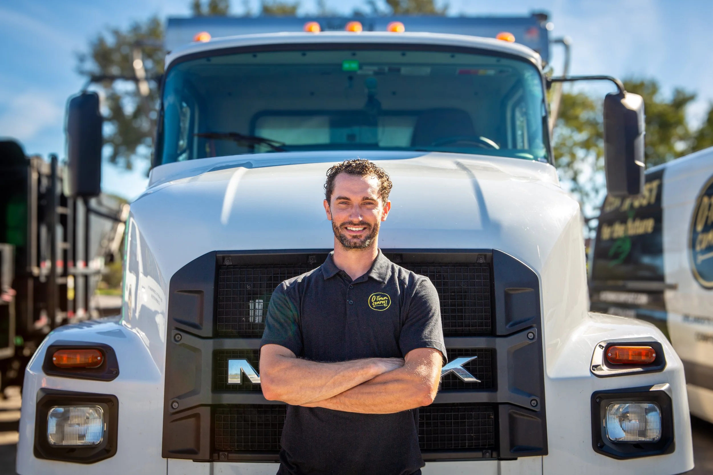 A man with curly hair and a beard standing with his arms crossed in front of a large white truck, smiling, with clear blue skies and trees in the background.