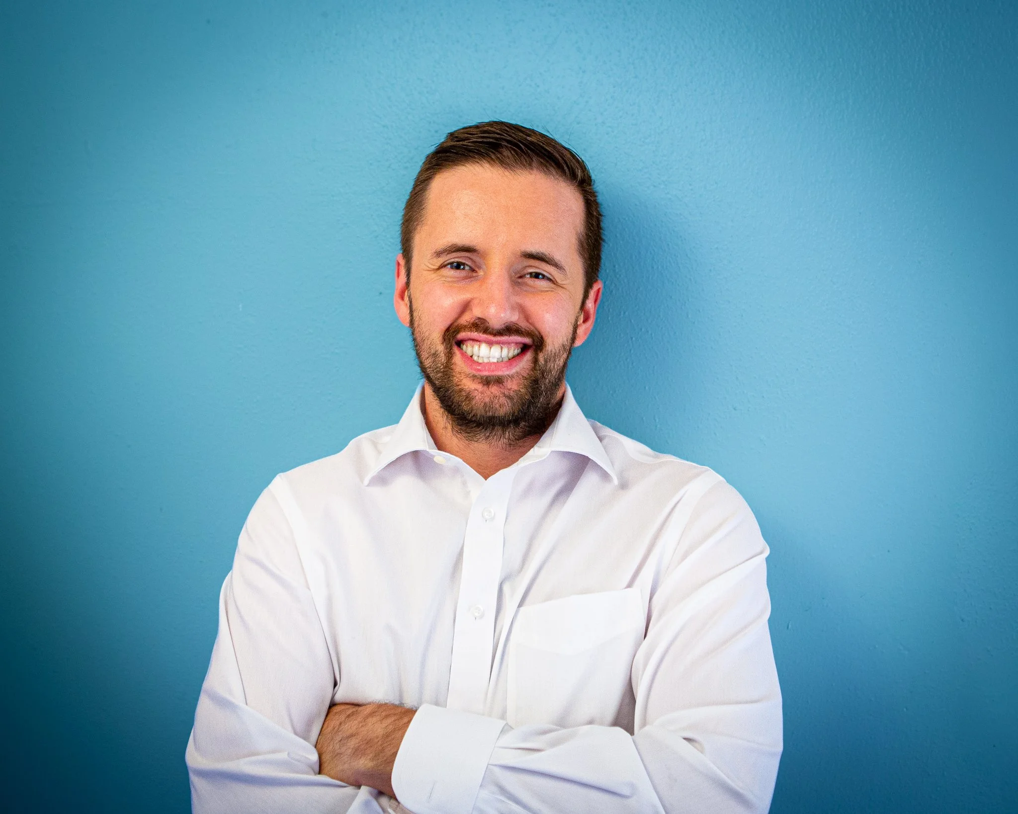A smiling man with dark hair and a beard standing against a bright blue wall, wearing a white shirt with arms crossed.