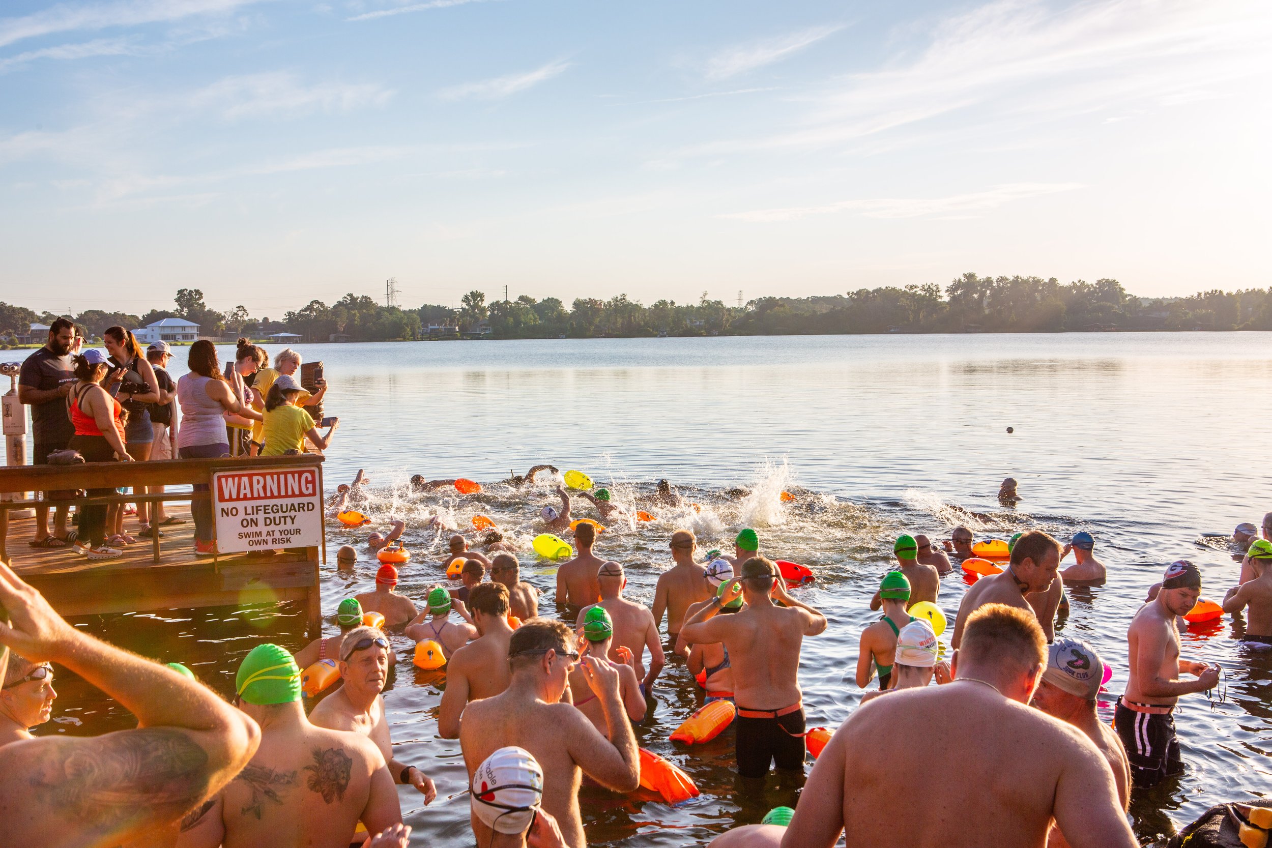 Swimmers wearing colorful swim caps and goggles entering a lake for a swim race, with spectators on a dock watching. The scene is set during sunny weather with a scenic background of trees and houses across the water.
