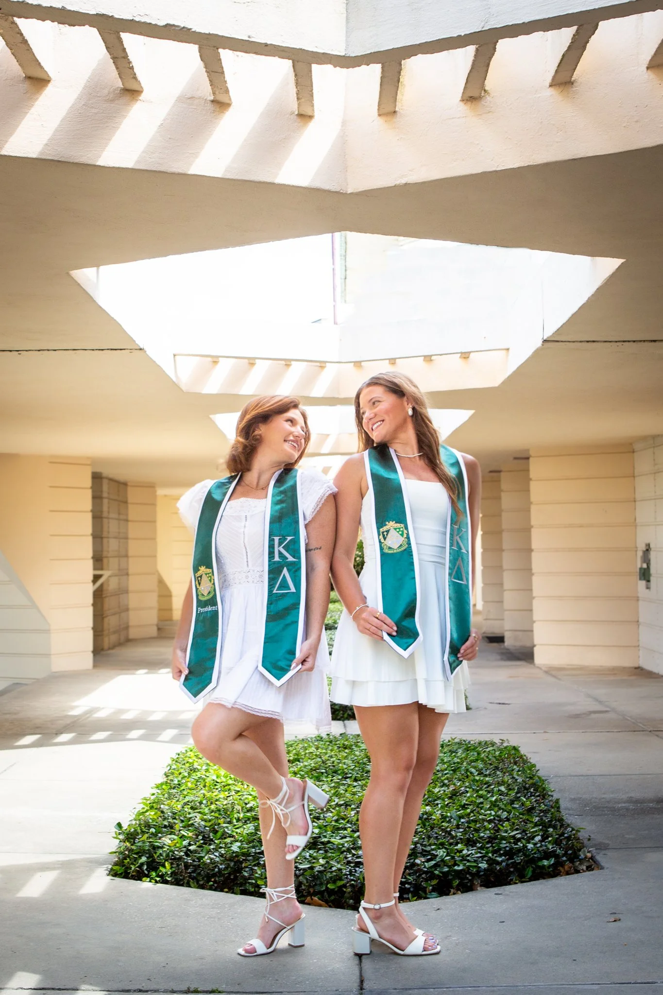 Two women wearing white dresses and green sashes with Greek letters, standing outdoors under a modern concrete architectural structure with geometric cut-outs, smiling and looking at each other.