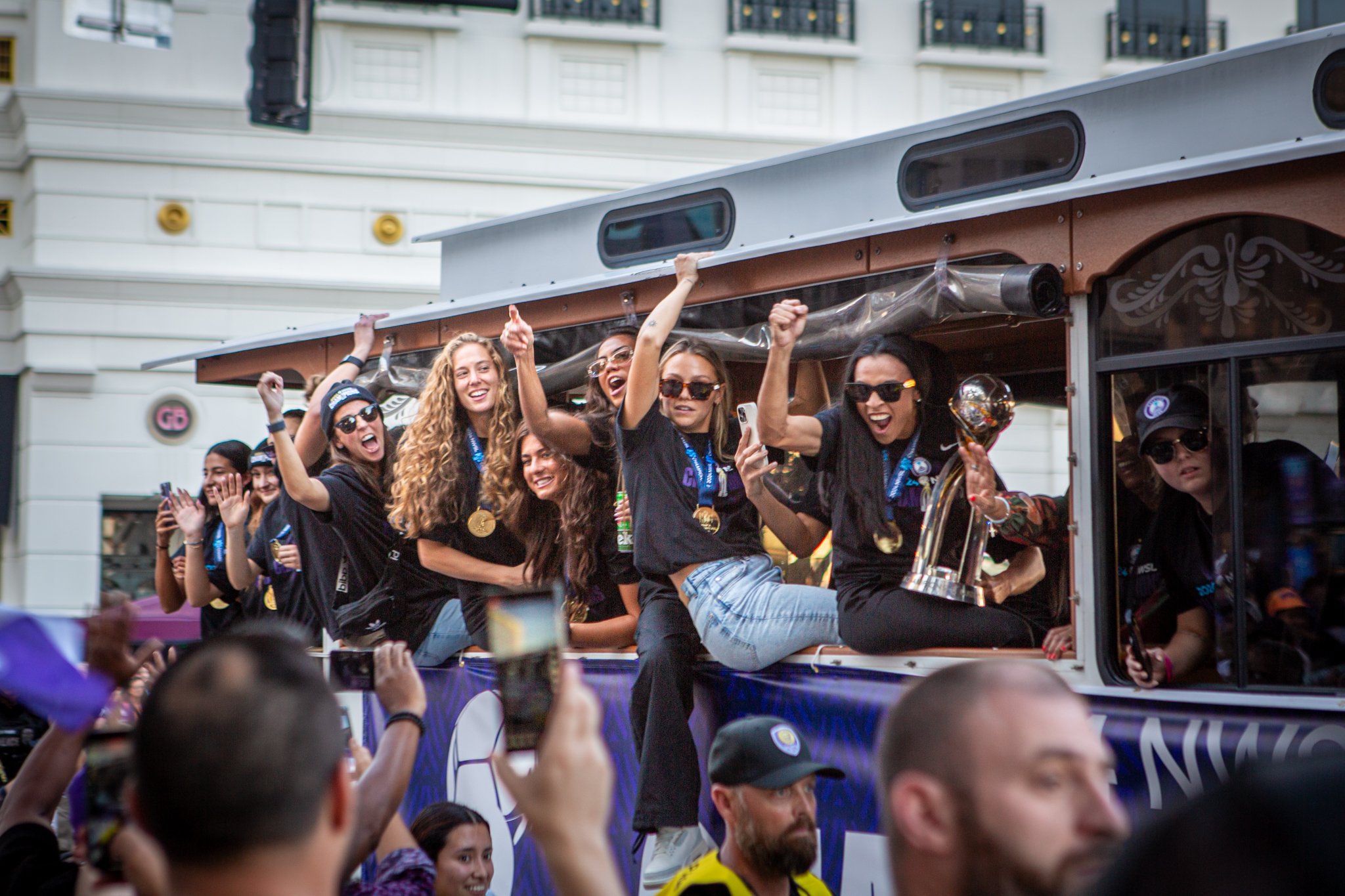 Group of people on a parade float celebrating with medals, some wearing sunglasses and black shirts, in a city street with buildings in the background.