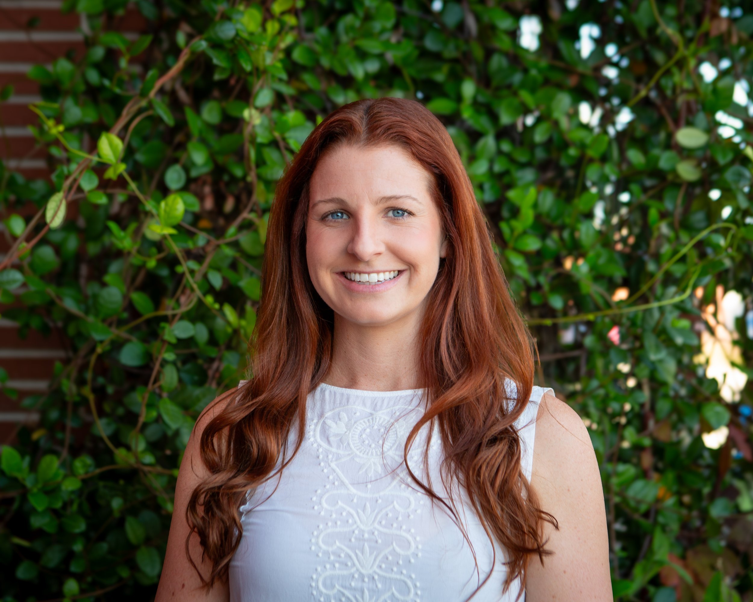 A woman with long red hair and blue eyes smiling outdoors in front of a green leafy bush.