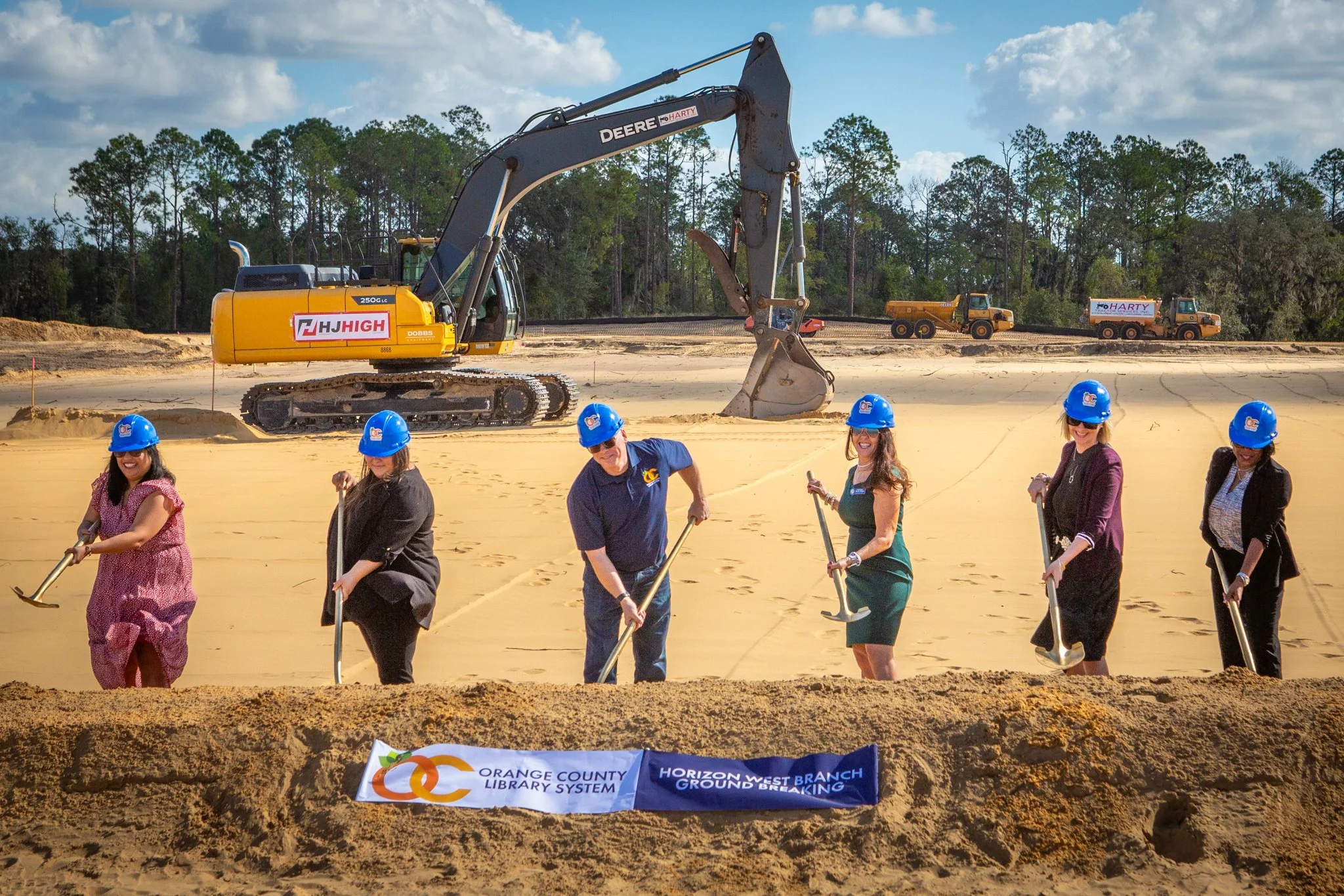 Group of people with blue hard hats planting a tree at a groundbreaking ceremony, with construction equipment and trees in the background.