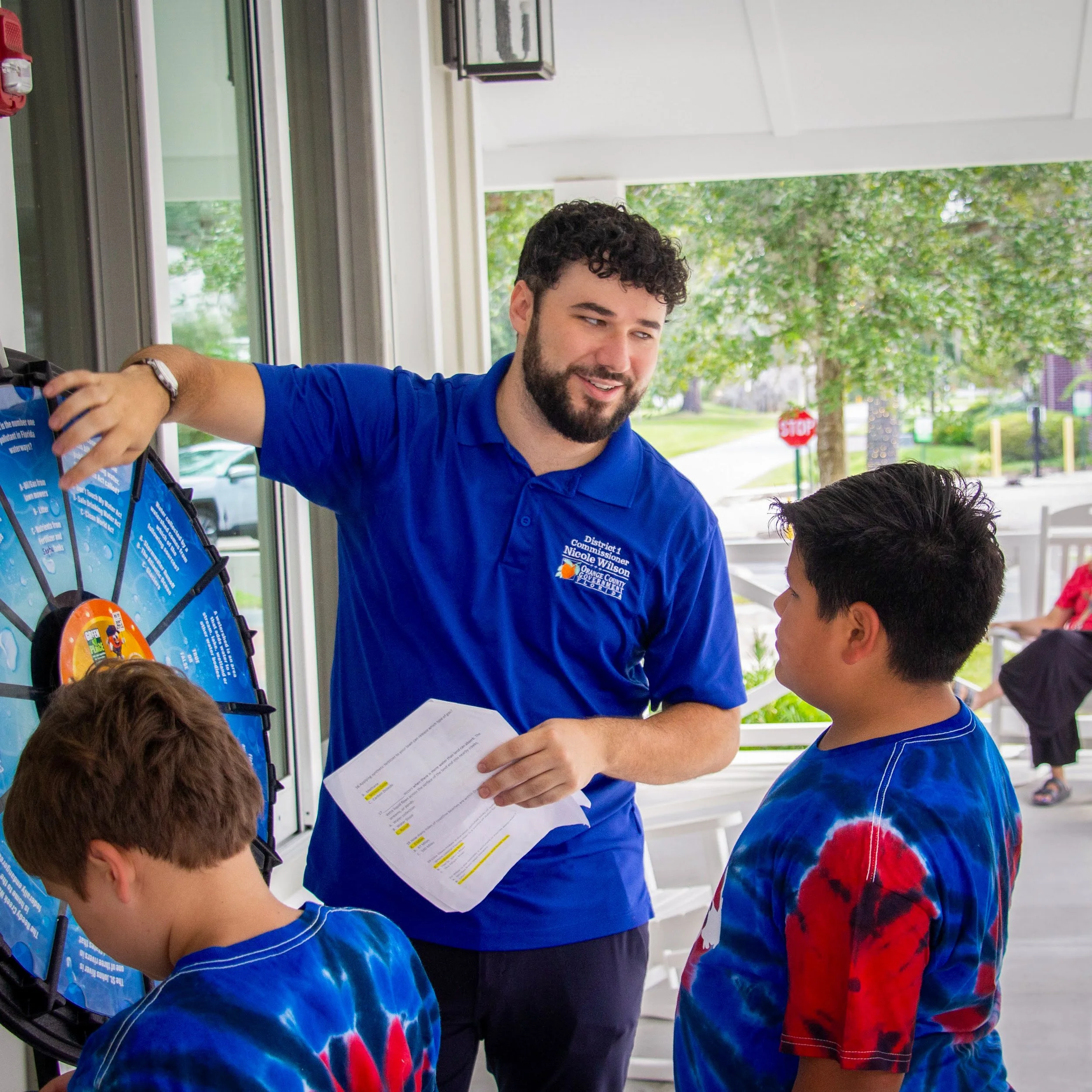 A man with curly hair and a beard wearing a blue polo shirt is handing out papers to a group of children, some with tie-dye shirts. The scene takes place outside a building with trees and a stop sign visible in the background.