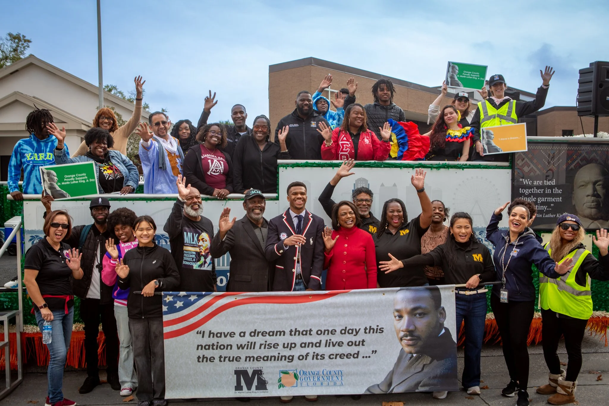 A diverse group of people at a parade or rally, some waving and smiling, holding signs and banners that promote Martin Luther King Jr. Day and support for the Black community, with a building and cloudy sky in the background.