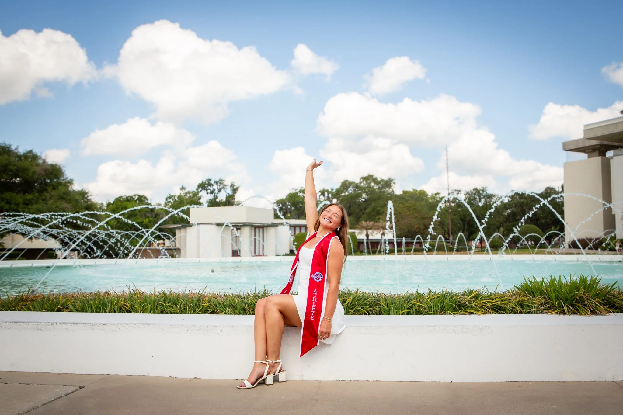 A young woman in a white dress with a red athletic sash sitting by a fountain, smiling and raising one arm, outdoors on a sunny day with a blue sky, white clouds, trees, and a building in the background.