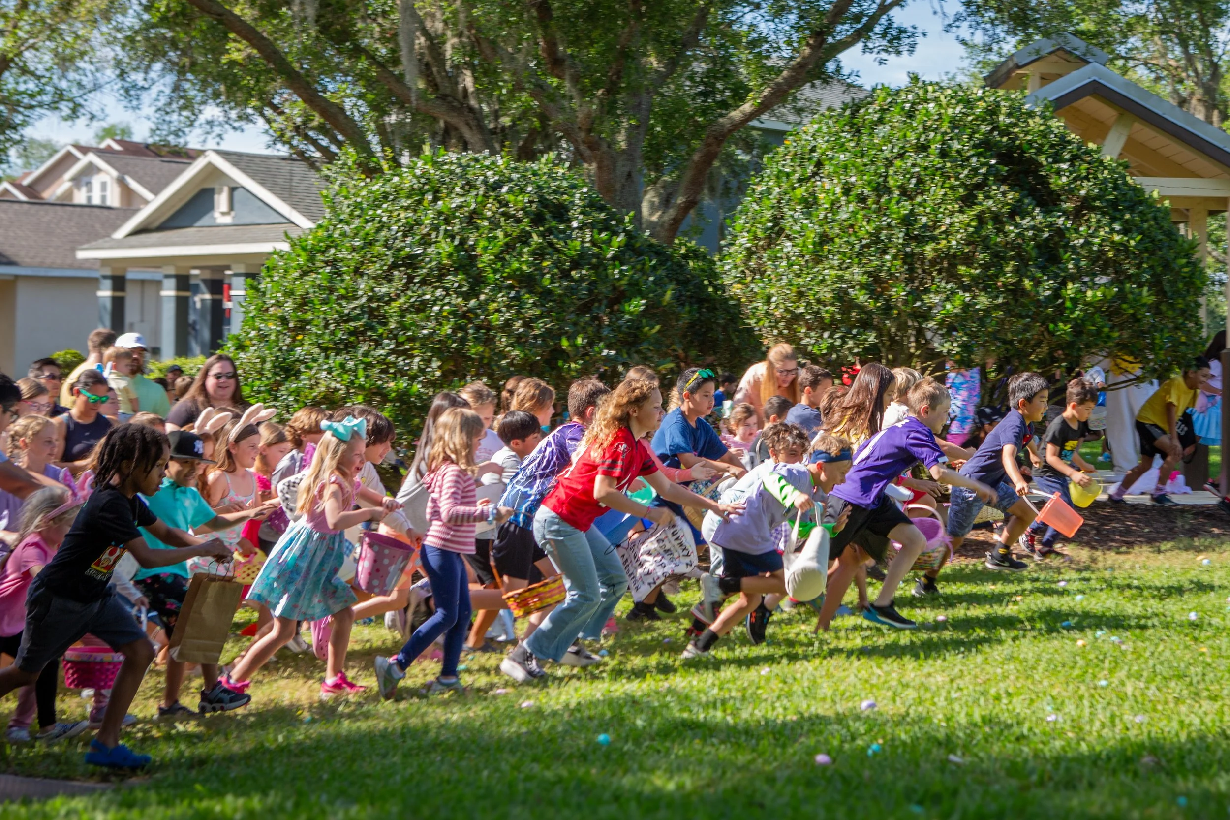 Children participating in an Easter egg hunt outdoors on a sunny day, running across a grassy area under trees with houses in the background.