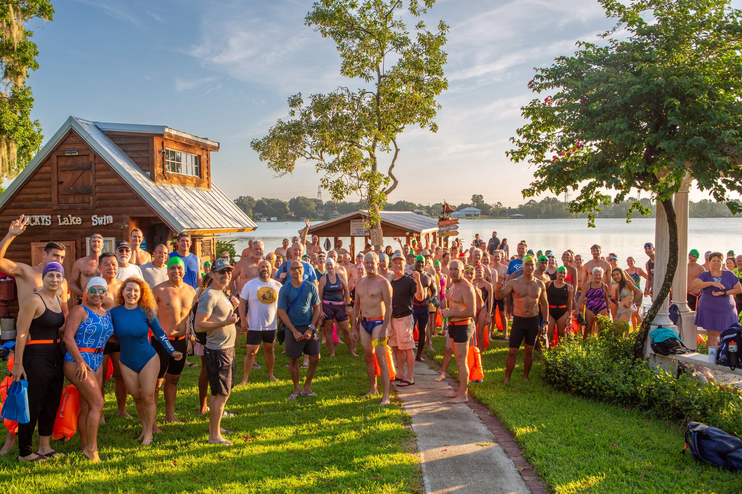 Group of people gathered outdoors at a swim event by the lake, many in swimsuits, some wearing swim caps, with trees, water, and a small wooden building in the background.