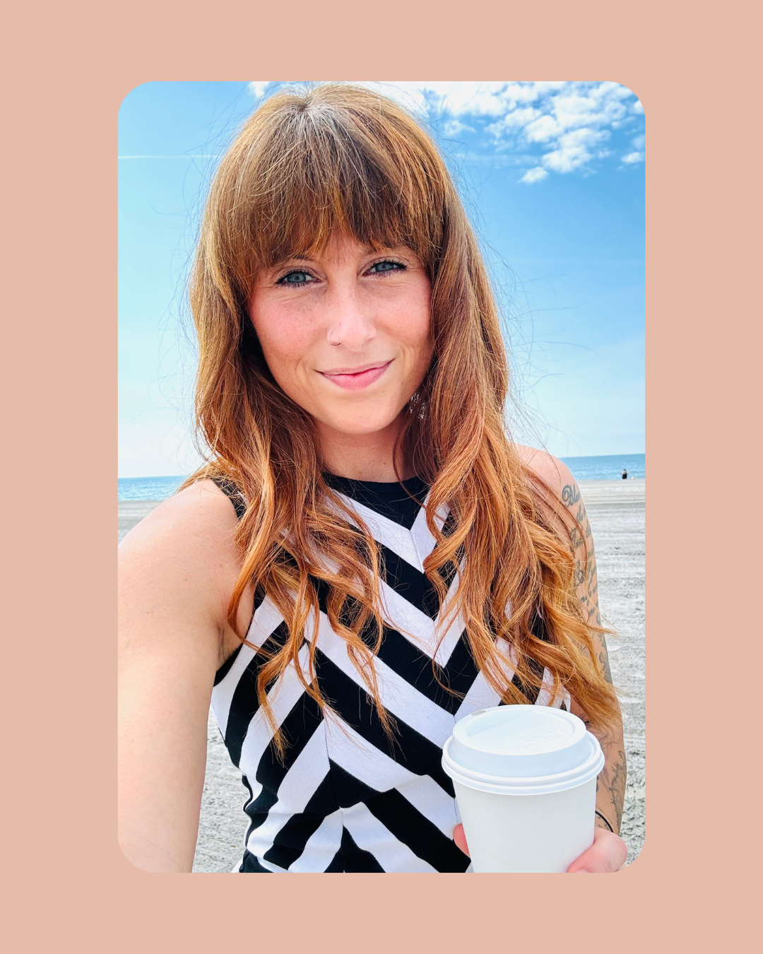 A woman with red hair, blue eyes, and a nose ring, smiling and holding a white coffee cup on a beach with a clear sky and ocean in the background.