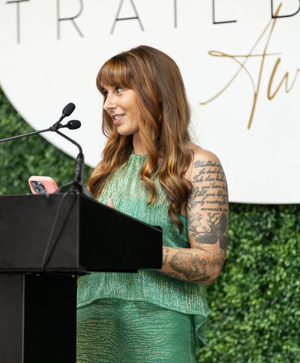 A woman with long red hair and tattoos on her left arm, wearing a sparkly green dress, stands at a black podium with microphones, looking to her left, in front of a white circular background with partially visible black and gold lettering and green foliage.