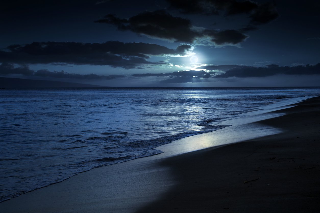 Nighttime view of the ocean with a full moon partially covered by clouds, reflecting on the water, near the sandy shoreline.