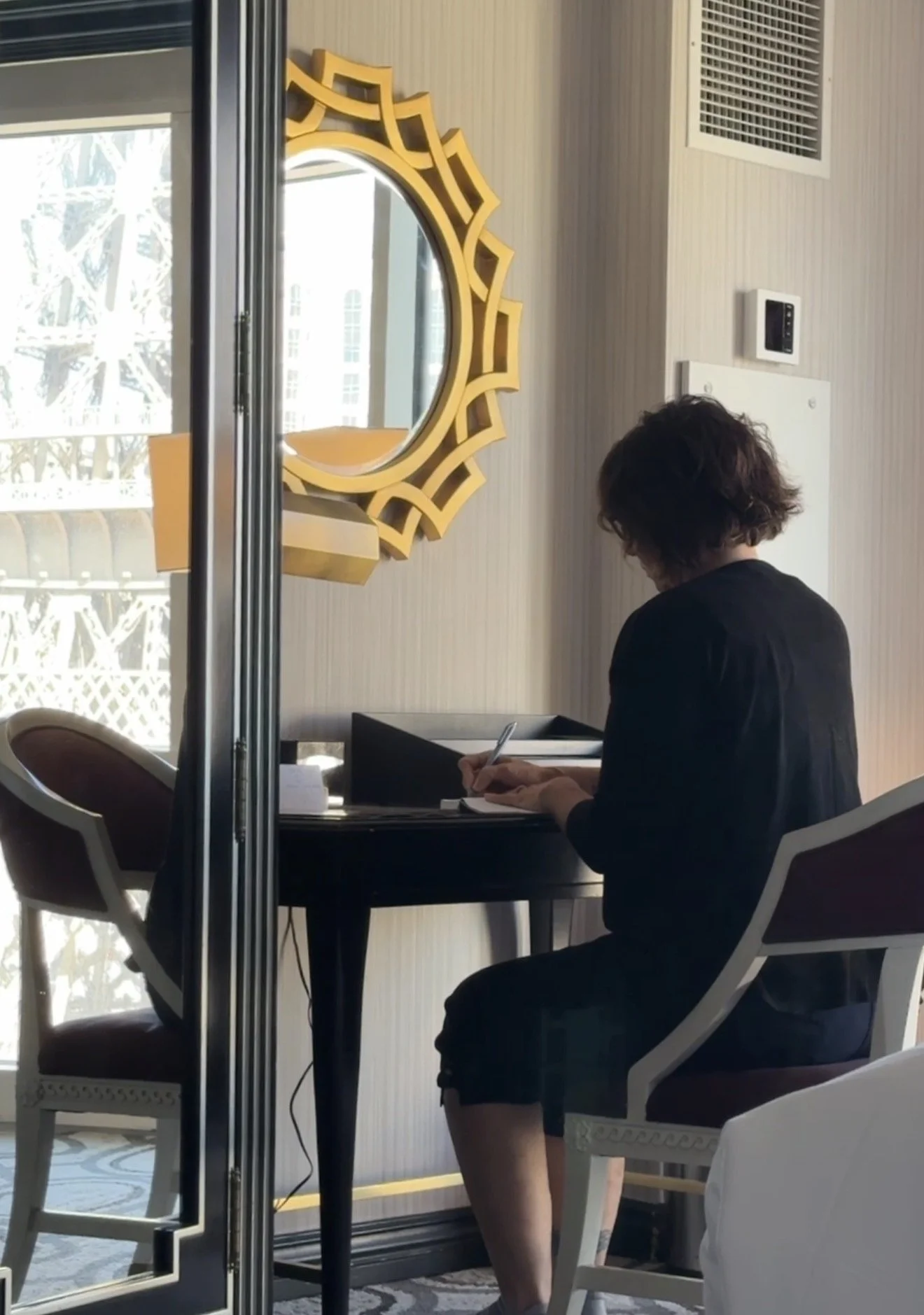 Woman sitting at a desk outdoors, writing on a notepad with a pen, beside a mirror with a decorative yellow frame, with a modern building visible through sliding glass doors.