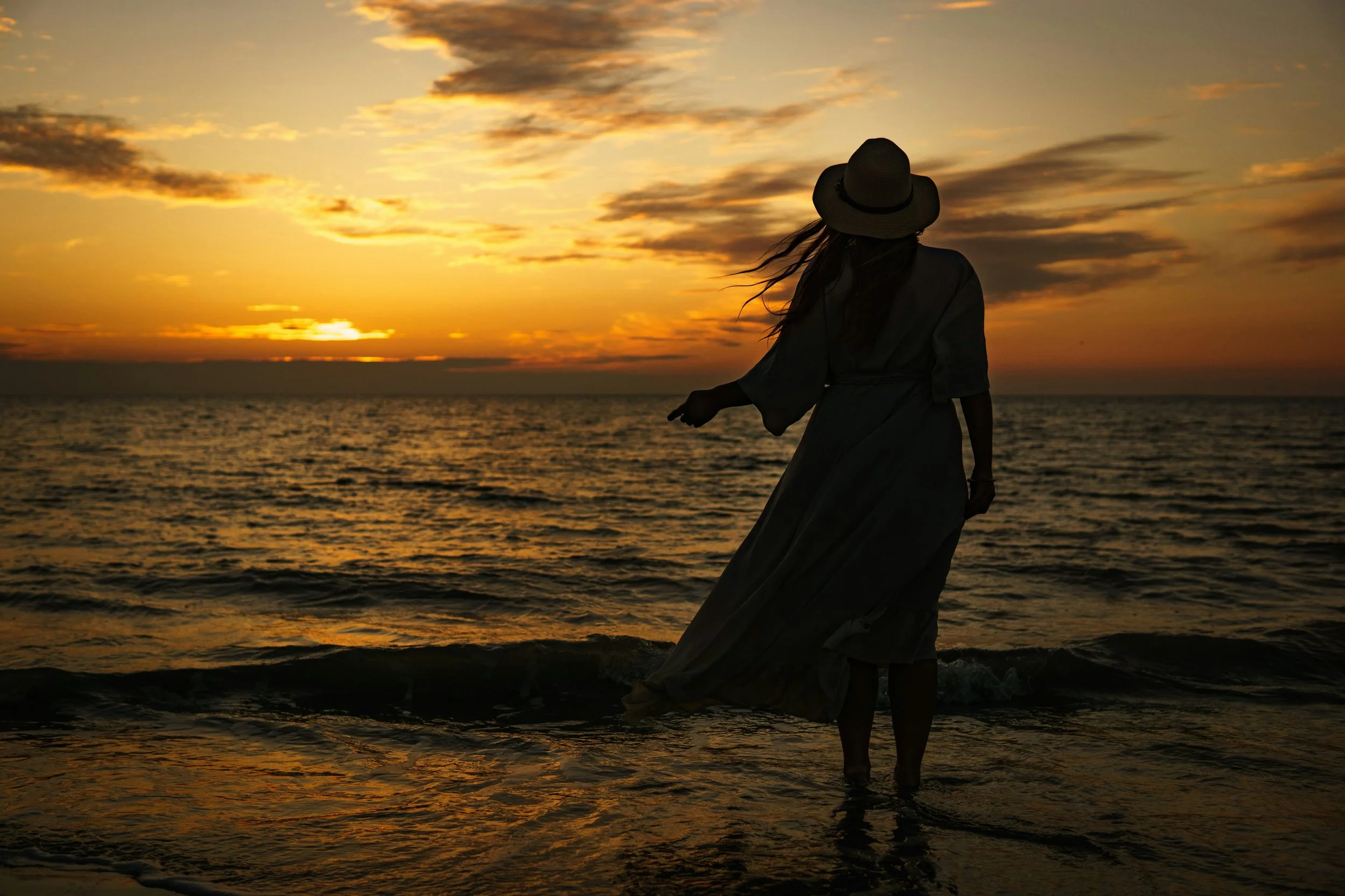 A woman wearing a hat and dress stands on the beach during sunset, facing the ocean and clouds with her hair blowing in the wind.