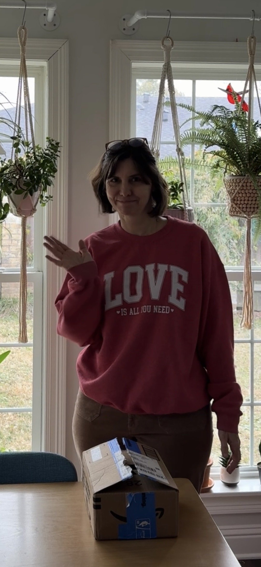A woman standing inside a house near a window with hanging plants. She is waving and smiling, wearing a red sweatshirt that says "LOVE IS ALL YOU NEED." There is a package on the table in front of her.