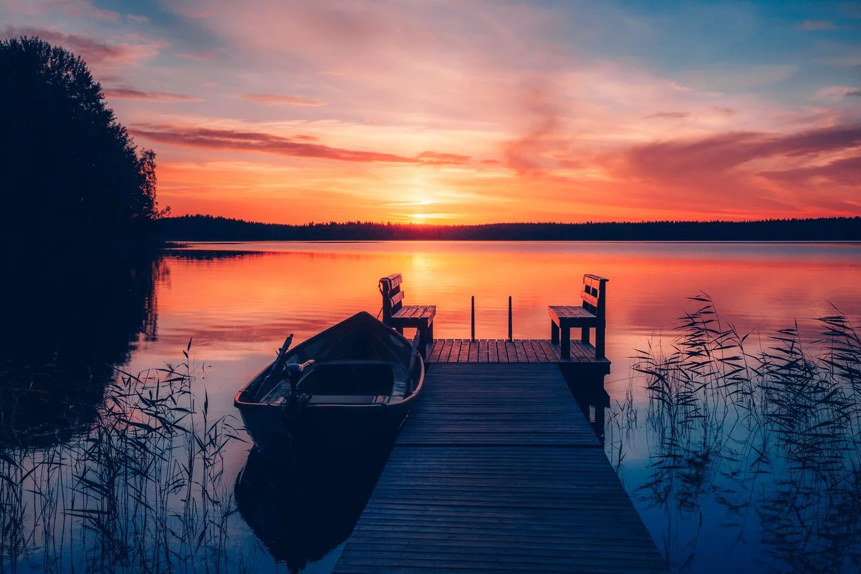 Sunset over a lake with a wooden dock and two benches, a rowboat moored to the side, and tall grasses in the foreground.